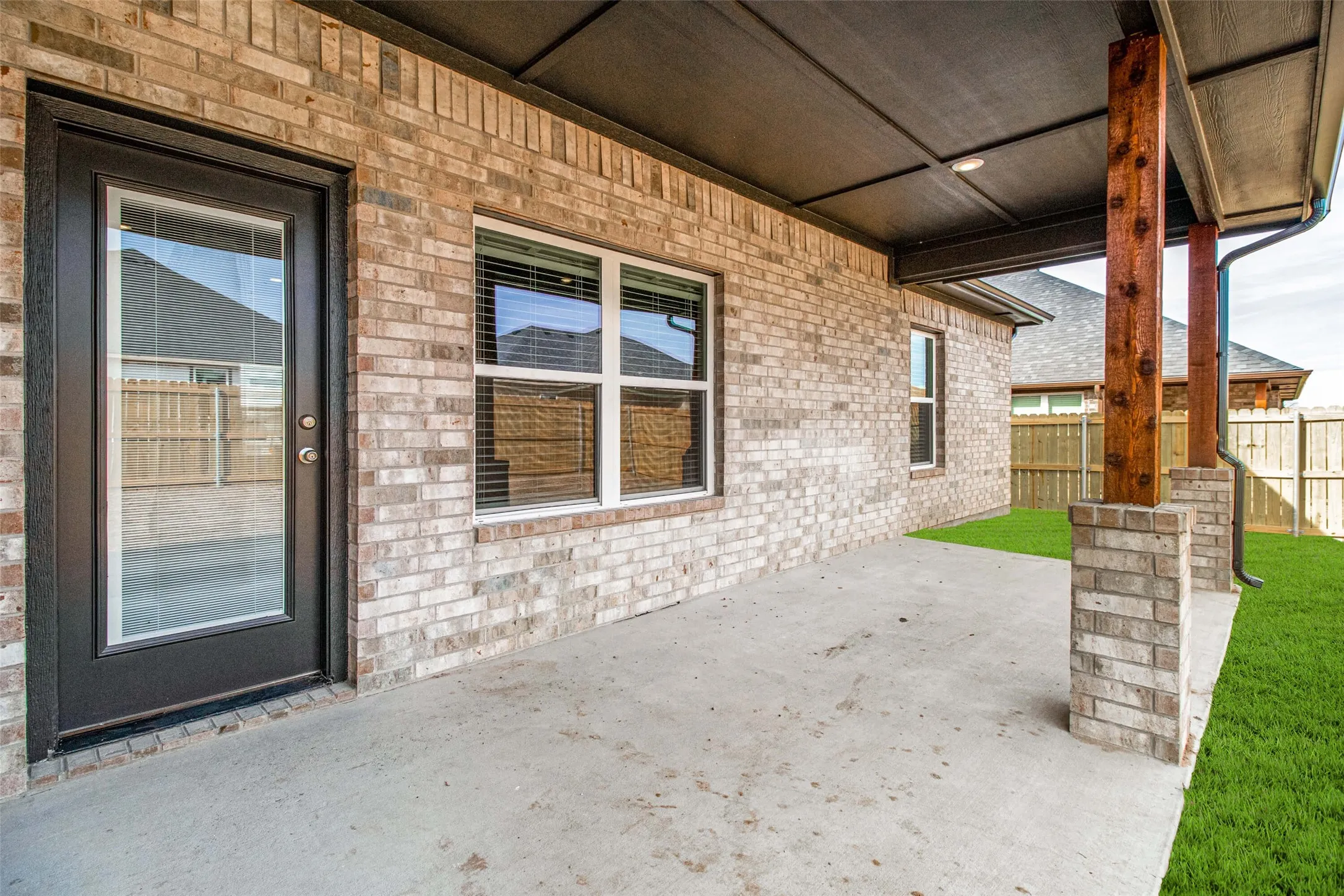 Covered patio featuring concrete flooring, a full-lite glass door, brick facade, and exposed wood posts