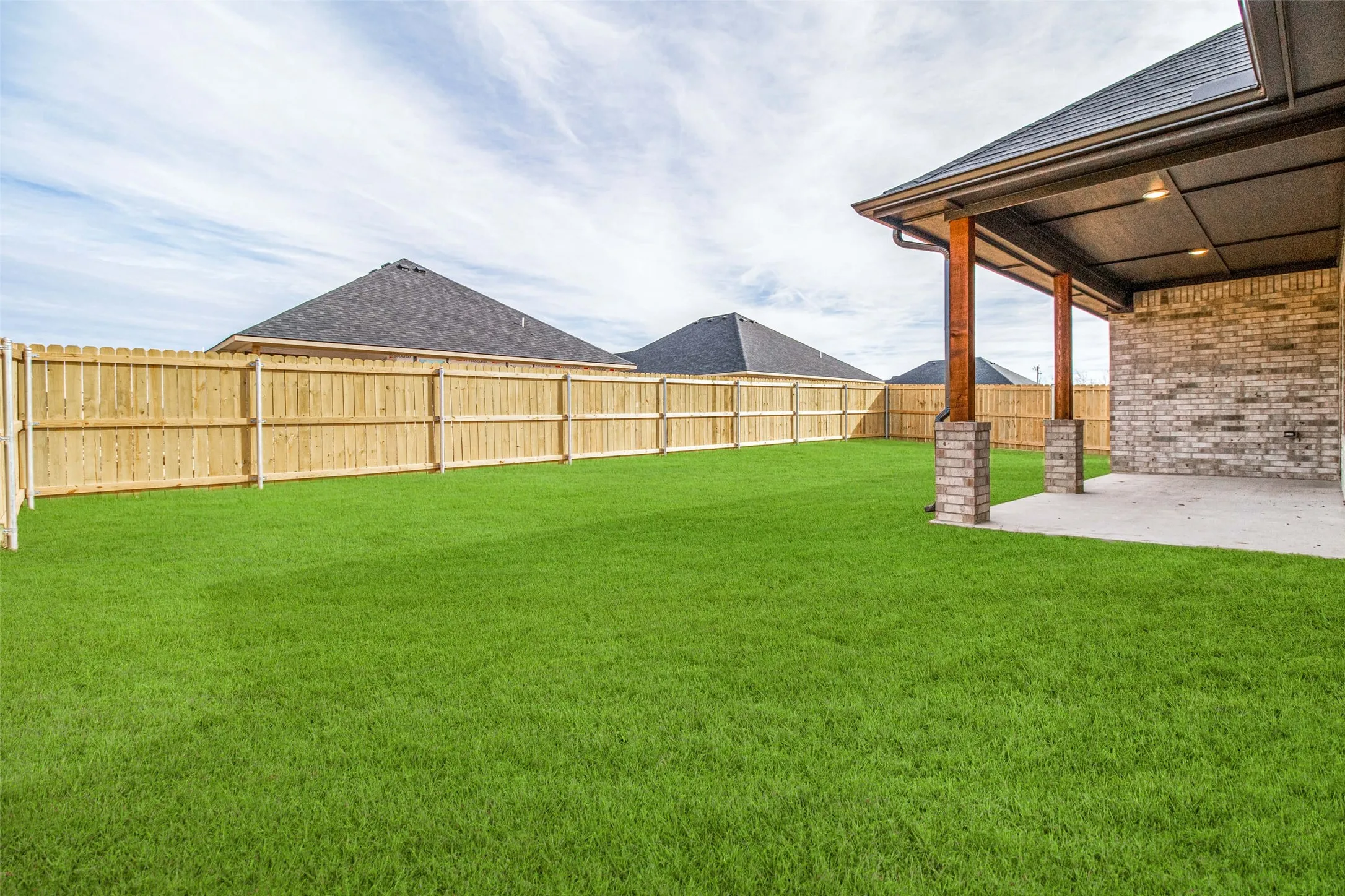 Expansive green lawn, wood privacy fence, covered concrete patio, brick exterior, and wood columns with stone bases