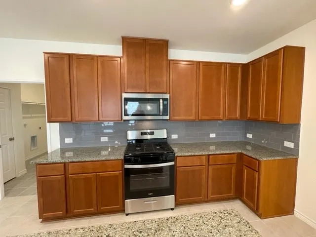 Wood-finish cabinetry, granite-style countertops, grey subway tile backsplash, stainless steel appliances, and tiled flooring