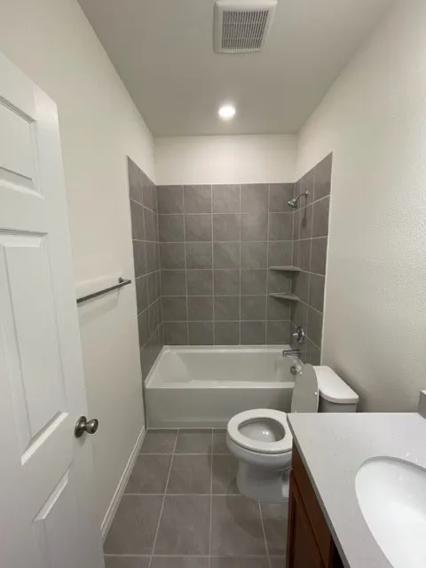 Bathtub with tile surround, built-in shelving, gray floor tiling, and a vanity with an integrated sink