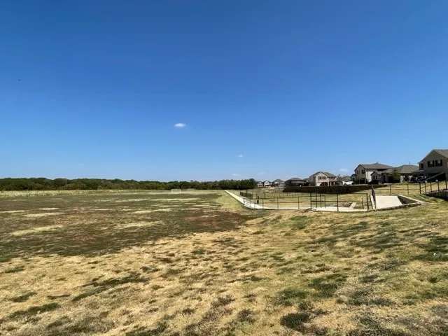 Expansive open field with a treeline backdrop, concrete pathways, and black metal fencing