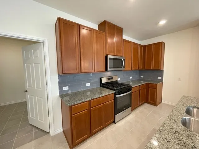 Kitchen featuring wood cabinetry, stainless steel appliances, granite countertops, a tiled backsplash, and ceramic tile flooring