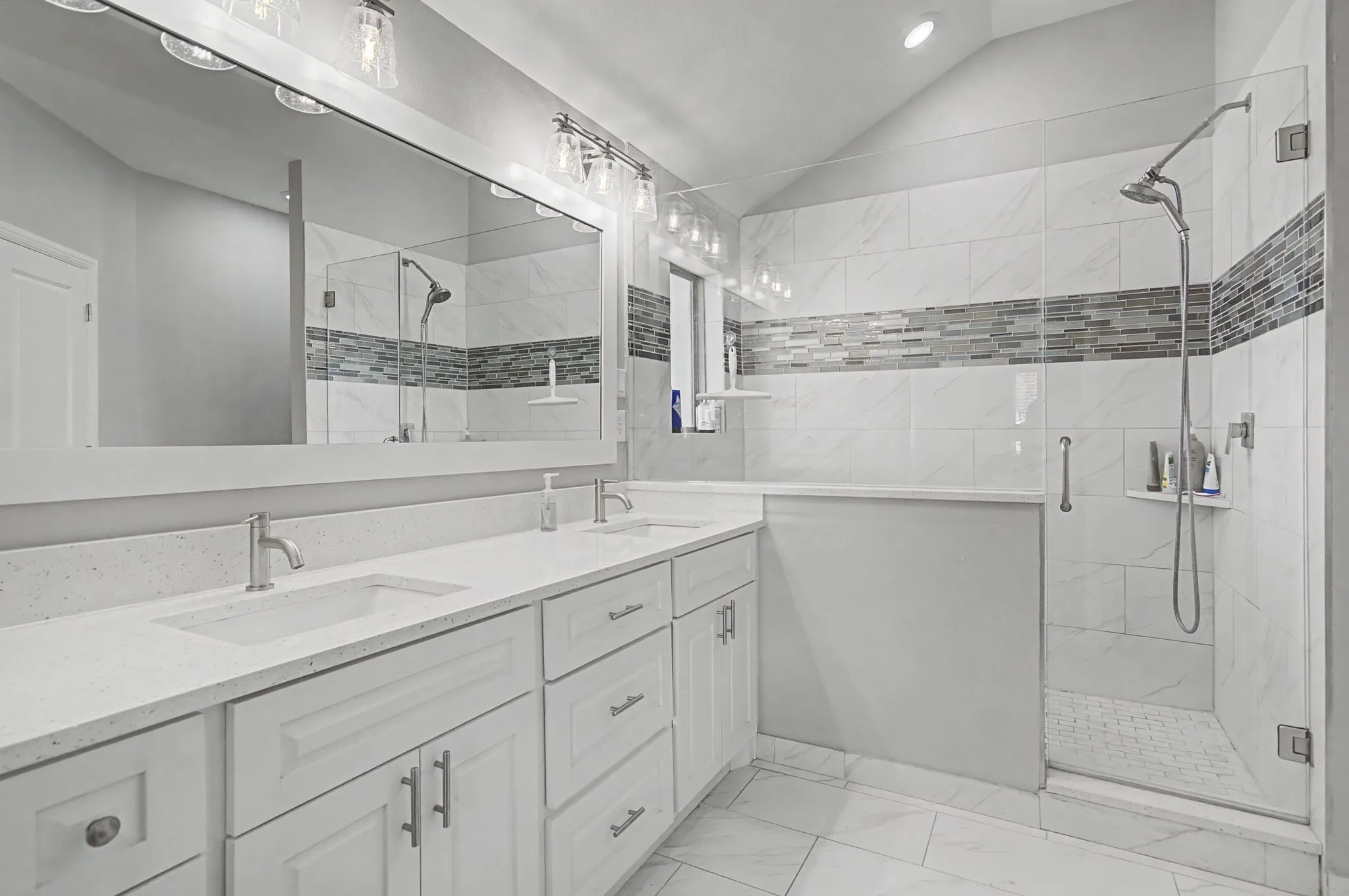 Dual vanity with integrated sinks and white countertops, extensive wall-mounted mirror, brushed nickel fixtures, and a glass-enclosed shower featuring a tiled accent band