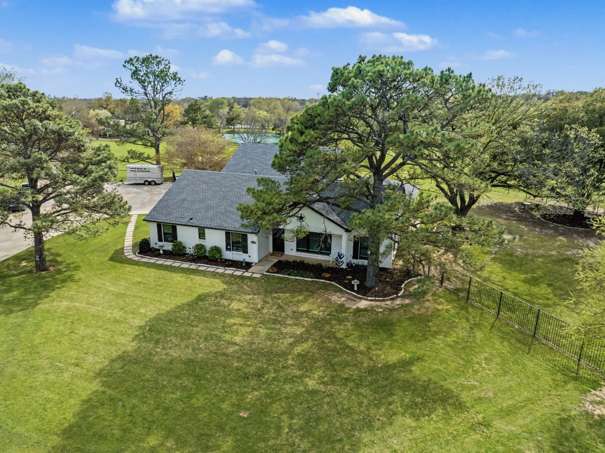 White exterior with dark shutters, dark roof, established trees, landscaped beds, and a perimeter fence