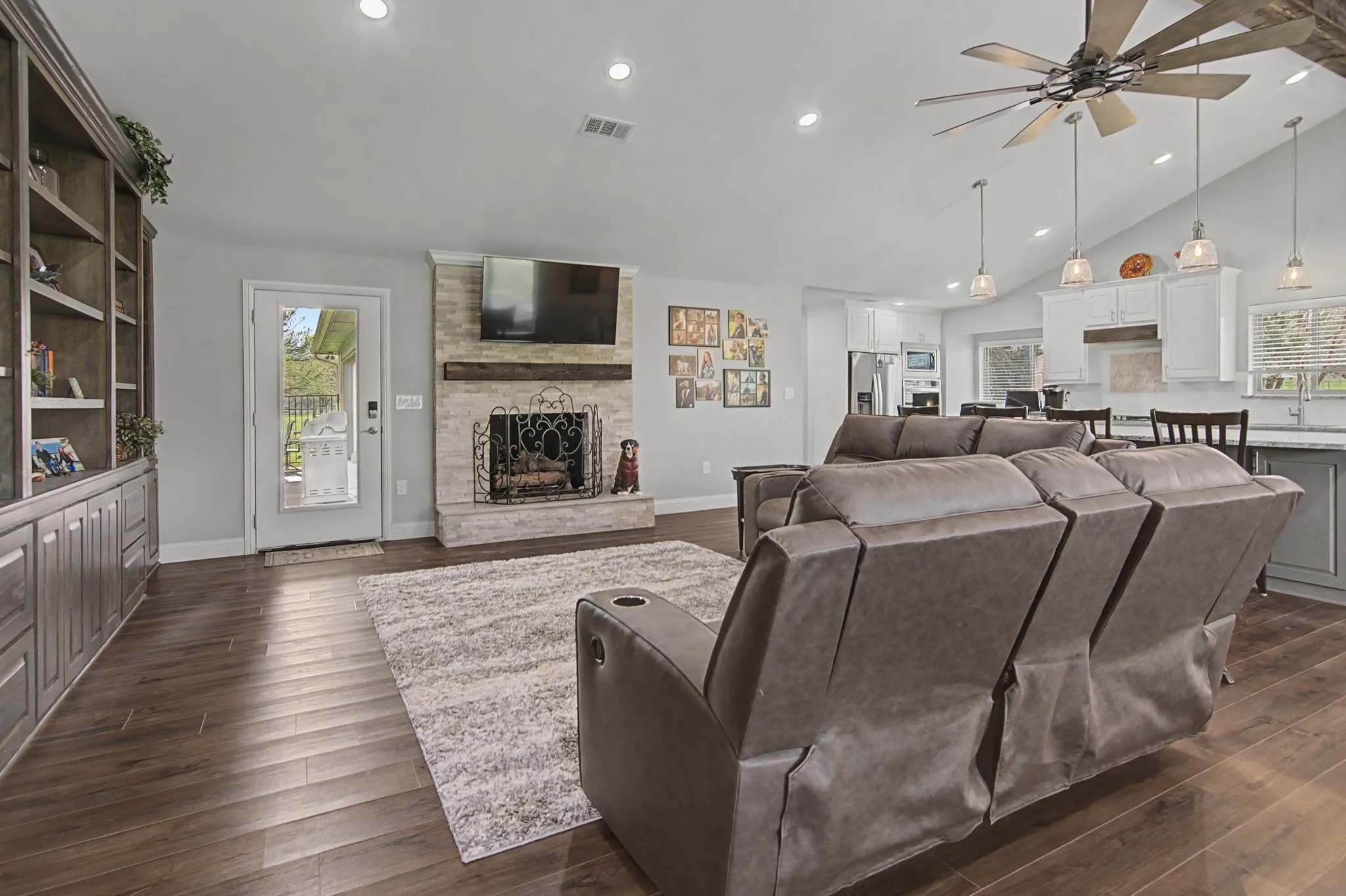 Open-concept living area featuring wood-finish flooring, a stone-faced fireplace with a wood mantle, and a vaulted ceiling with a large ceiling fan