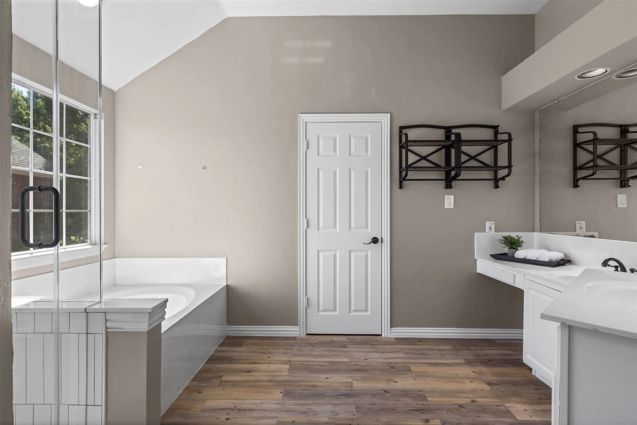 Soaking tub with a tiled surround, glass-enclosed shower, wood-finish flooring, dual vanity with undermount sinks, and recessed lighting