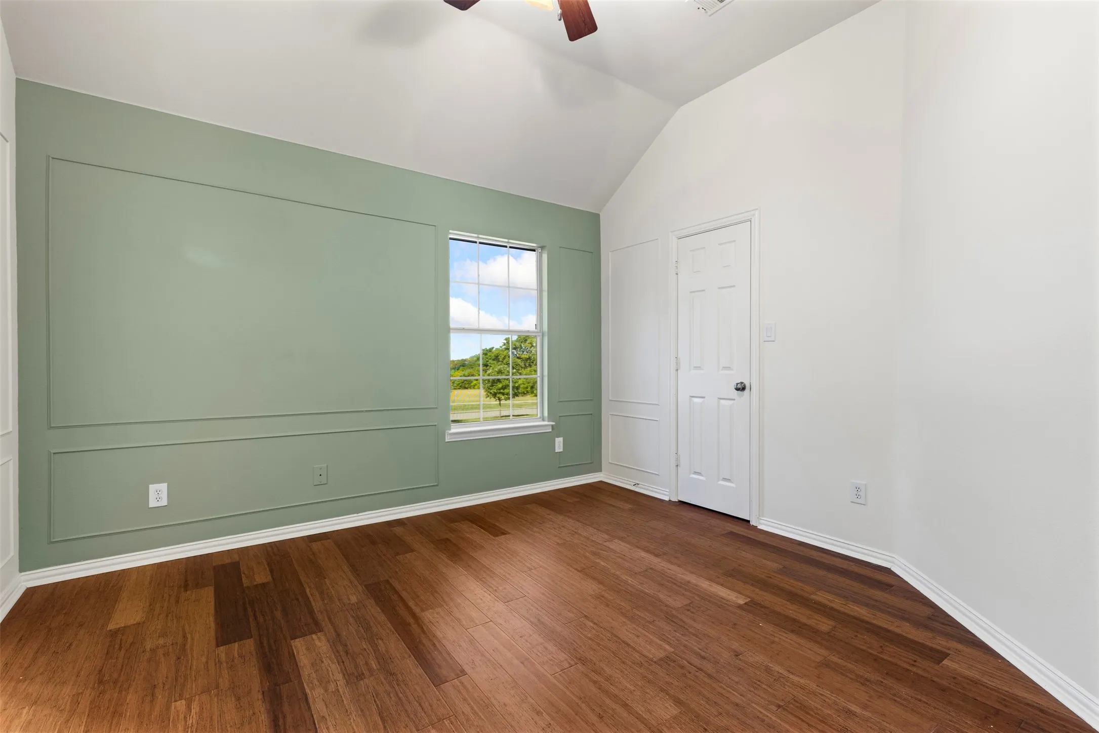 Vaulted ceiling, wood-finish flooring, accent wall with decorative trim, paneled door, ceiling fan