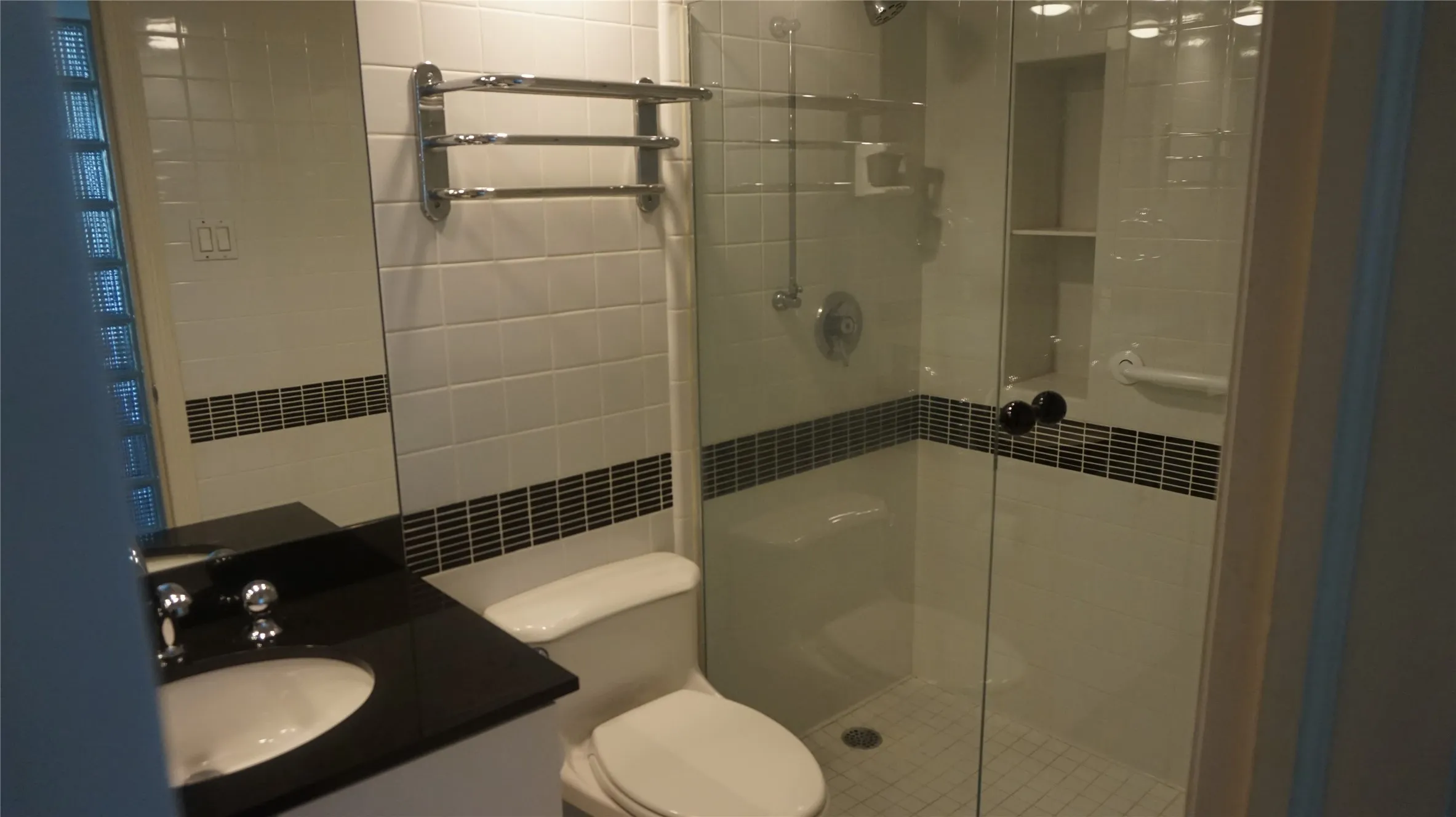 Bathroom featuring a glass-enclosed shower, built-in shower shelving, a dark countertop vanity with an integrated sink, and white subway tile with a decorative accent band