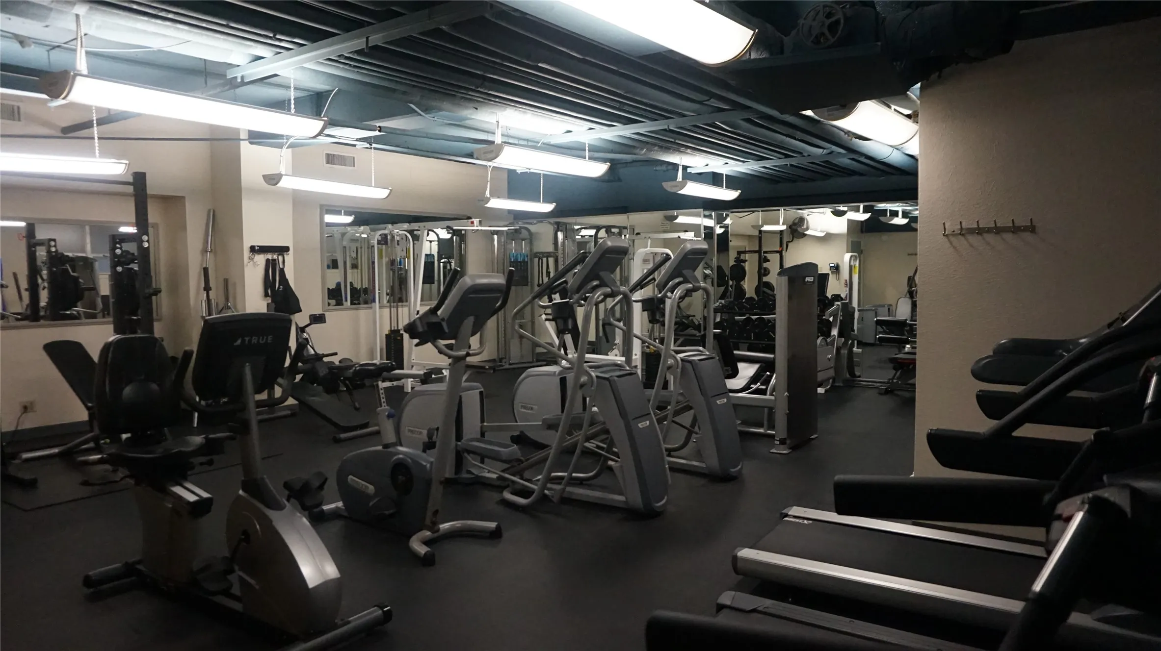 Fitness facility with a rubberized floor, mirrored wall, exposed ceiling, and fluorescent lighting