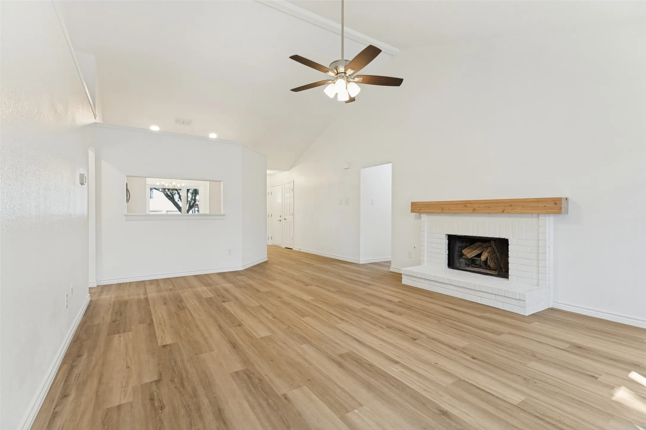 Vaulted ceiling with a ceiling fan, wood-finish flooring, white brick fireplace with a wood mantle, and an interior pass-through window