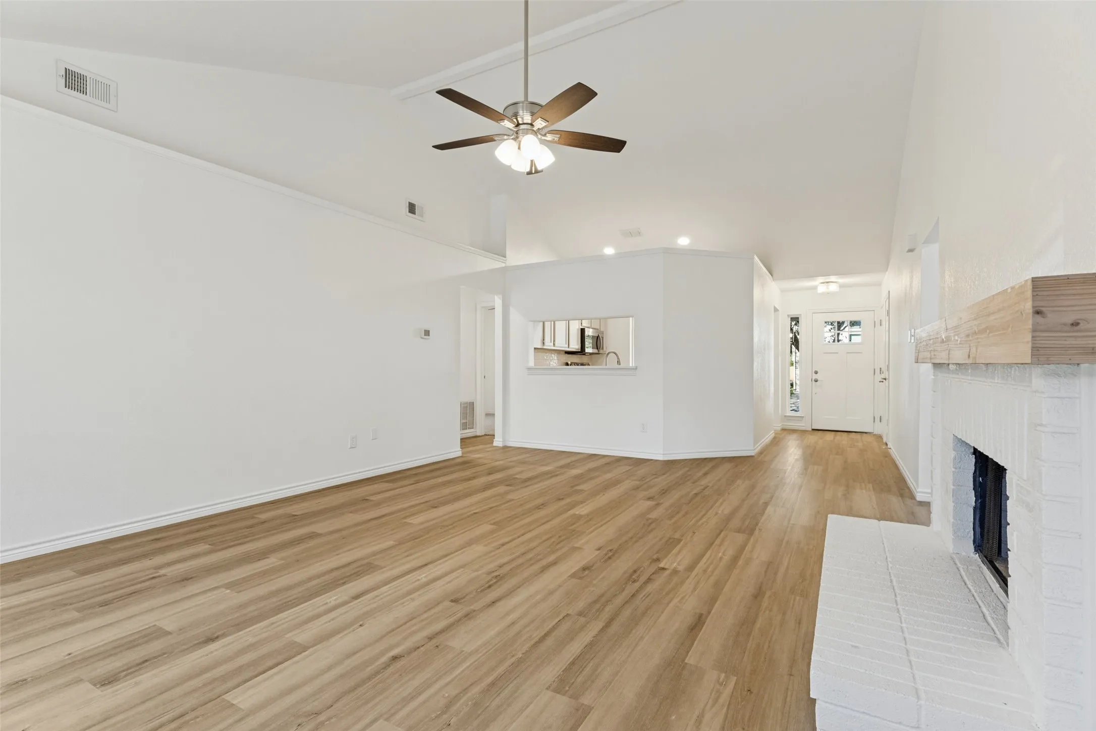Vaulted ceiling with ceiling fan, wood-finish flooring, white brick fireplace with wood mantel, built-in kitchen pass-through, and white paneled entry door with sidelights