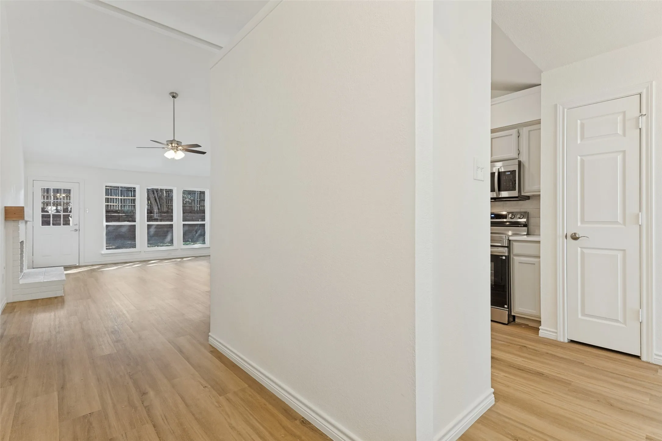 Vaulted ceiling with a ceiling fan, recessed windows, wood-finish flooring, white painted brick fireplace, and a white kitchen with stainless steel appliances