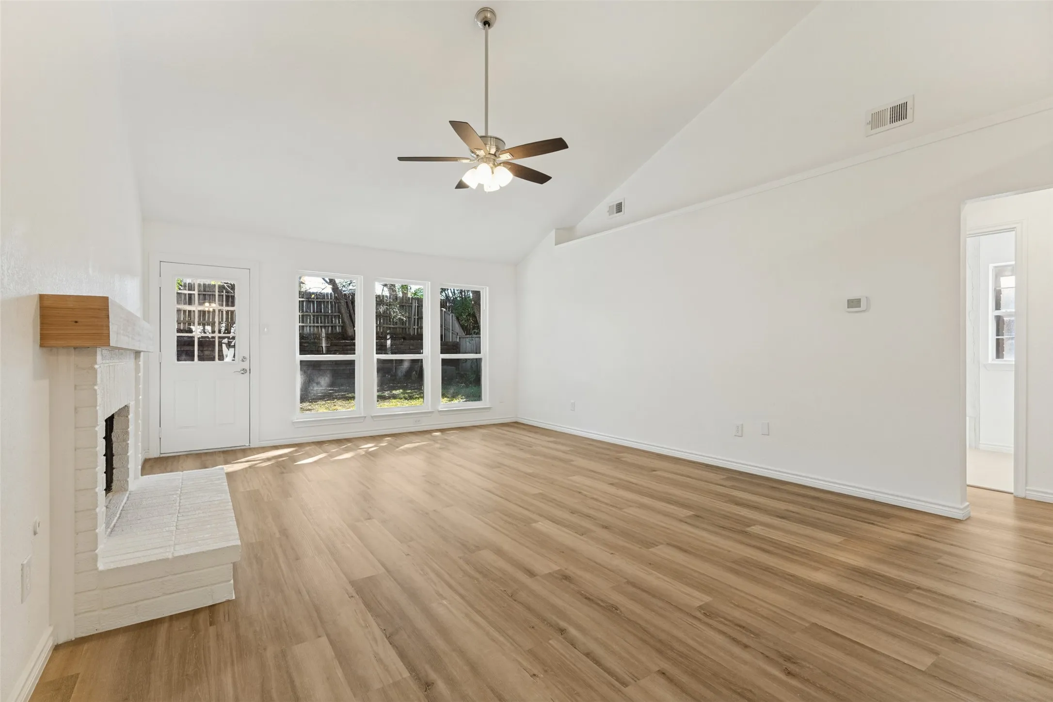 Vaulted ceiling with ceiling fan, wood-finish flooring, white brick fireplace with wood mantel, multiple windows, and a glass-paneled exterior door