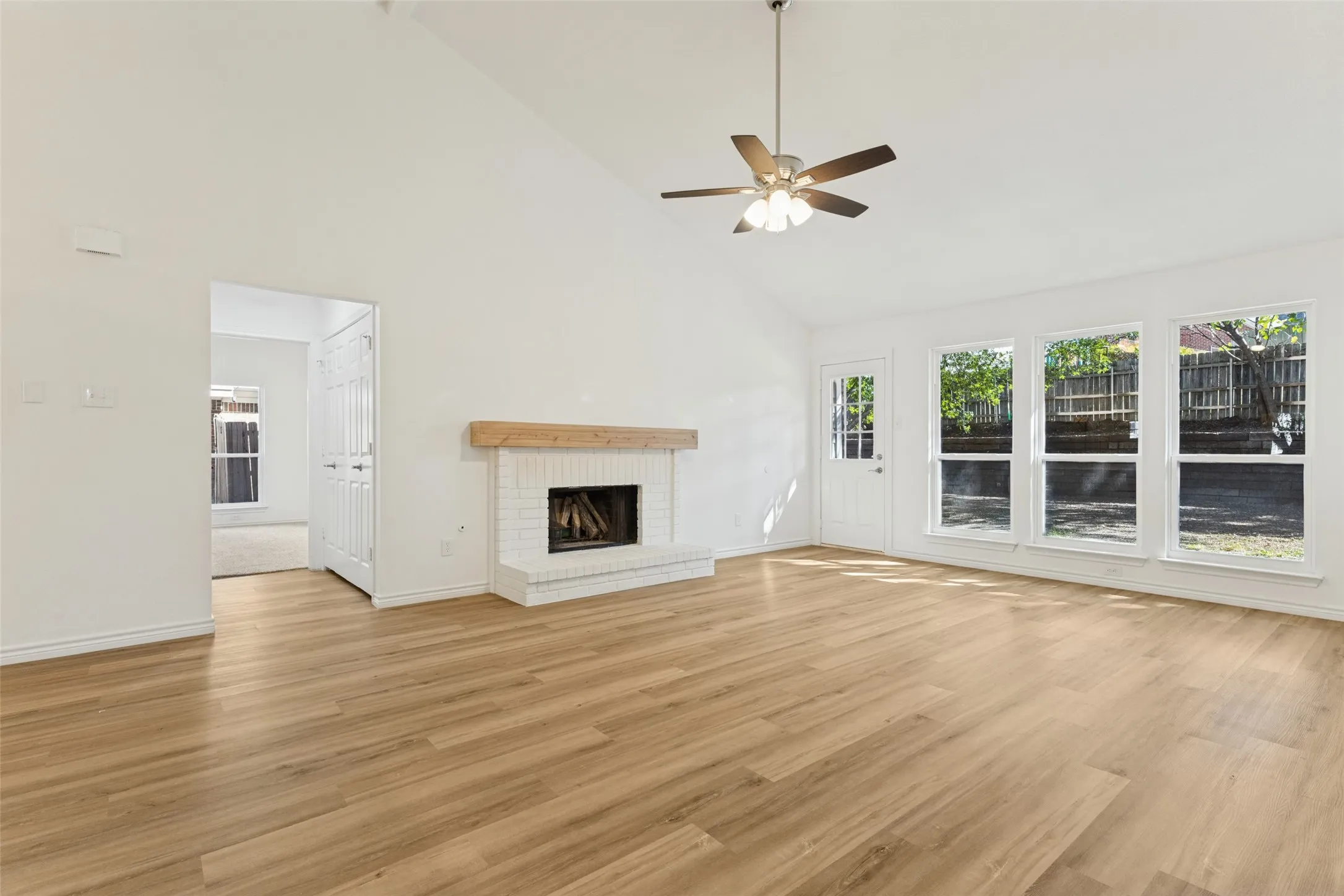 Vaulted ceiling, wood-finish flooring, white brick fireplace with wood mantel, ceiling fan, and multiple large windows