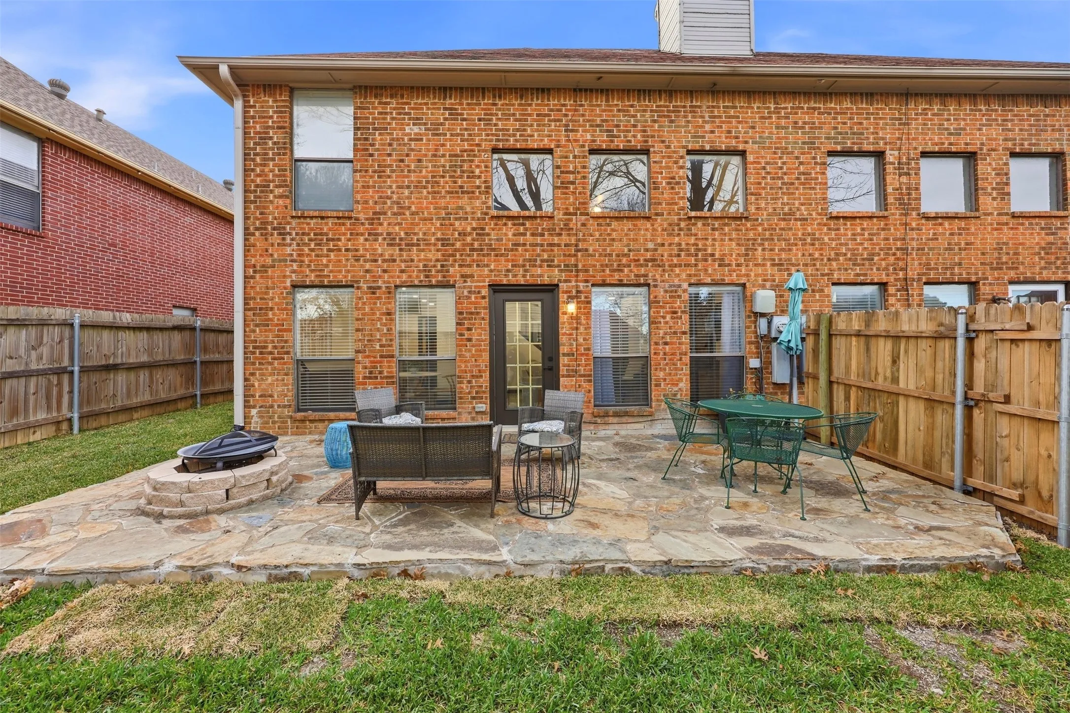 Red brick exterior with multiple windows, leading to a stone patio with a built-in fire pit