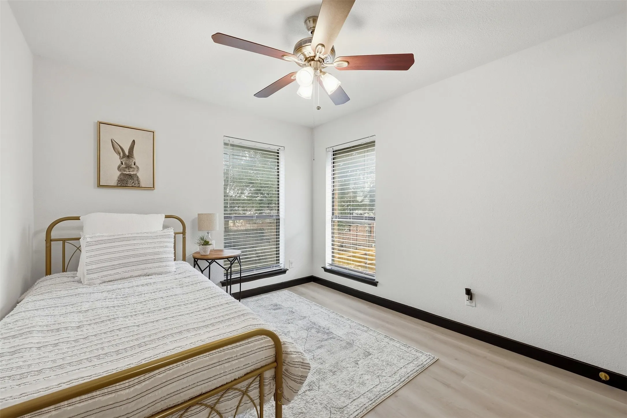 Light-toned wood-finish flooring, two large windows with blinds, ceiling fan with integrated lighting, and white painted walls