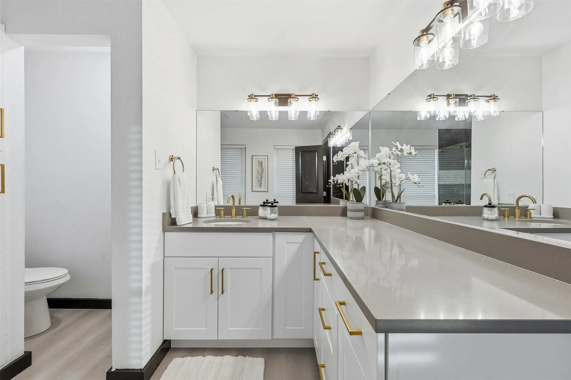 Dual vanity with undermount sinks, expansive countertop, white cabinetry with gold-tone hardware, and mirrored wall