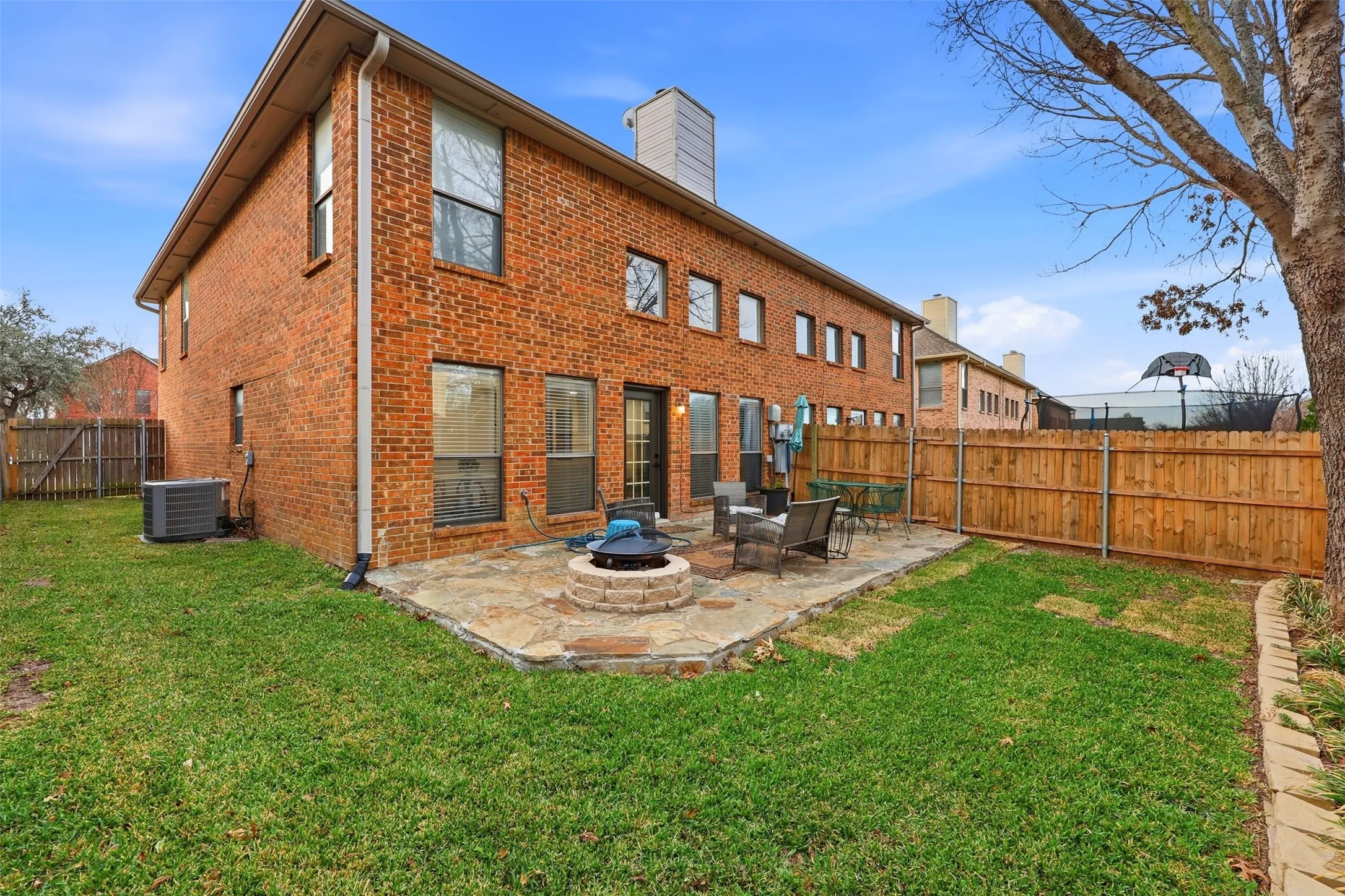 Brick exterior with multiple windows, featuring a flagstone patio with a built-in fire pit, surrounded by a wooden privacy fence and a grassy yard