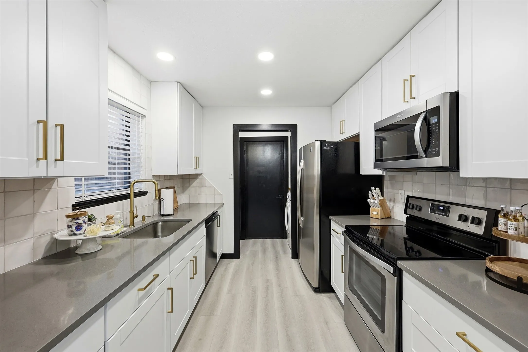 Galley kitchen featuring white cabinetry, gold-tone hardware, stainless steel appliances, a gold-tone faucet, and light wood-finish flooring