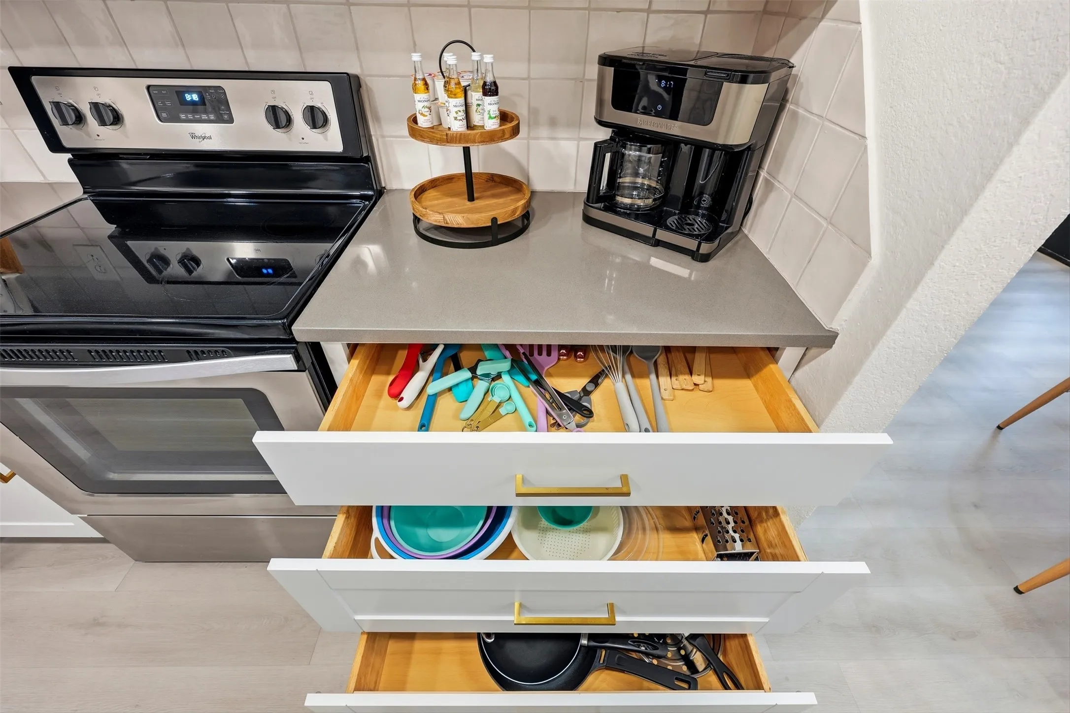 Stainless steel range, light-toned countertop, white cabinetry with gold-tone hardware, white tile backsplash, light wood-finish flooring