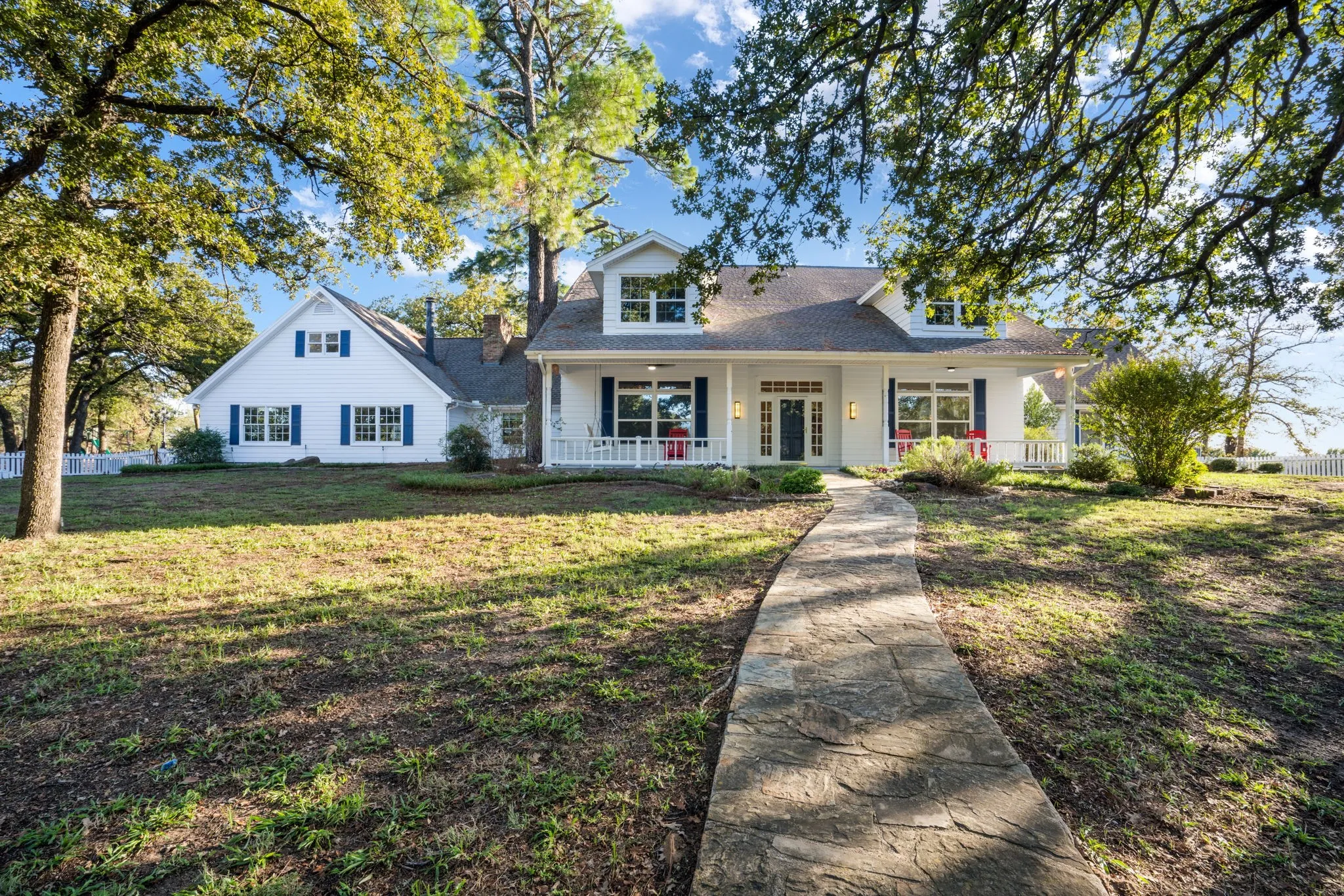 White exterior with blue shutters, a covered front porch, a stone pathway, a partial white picket fence, and mature trees