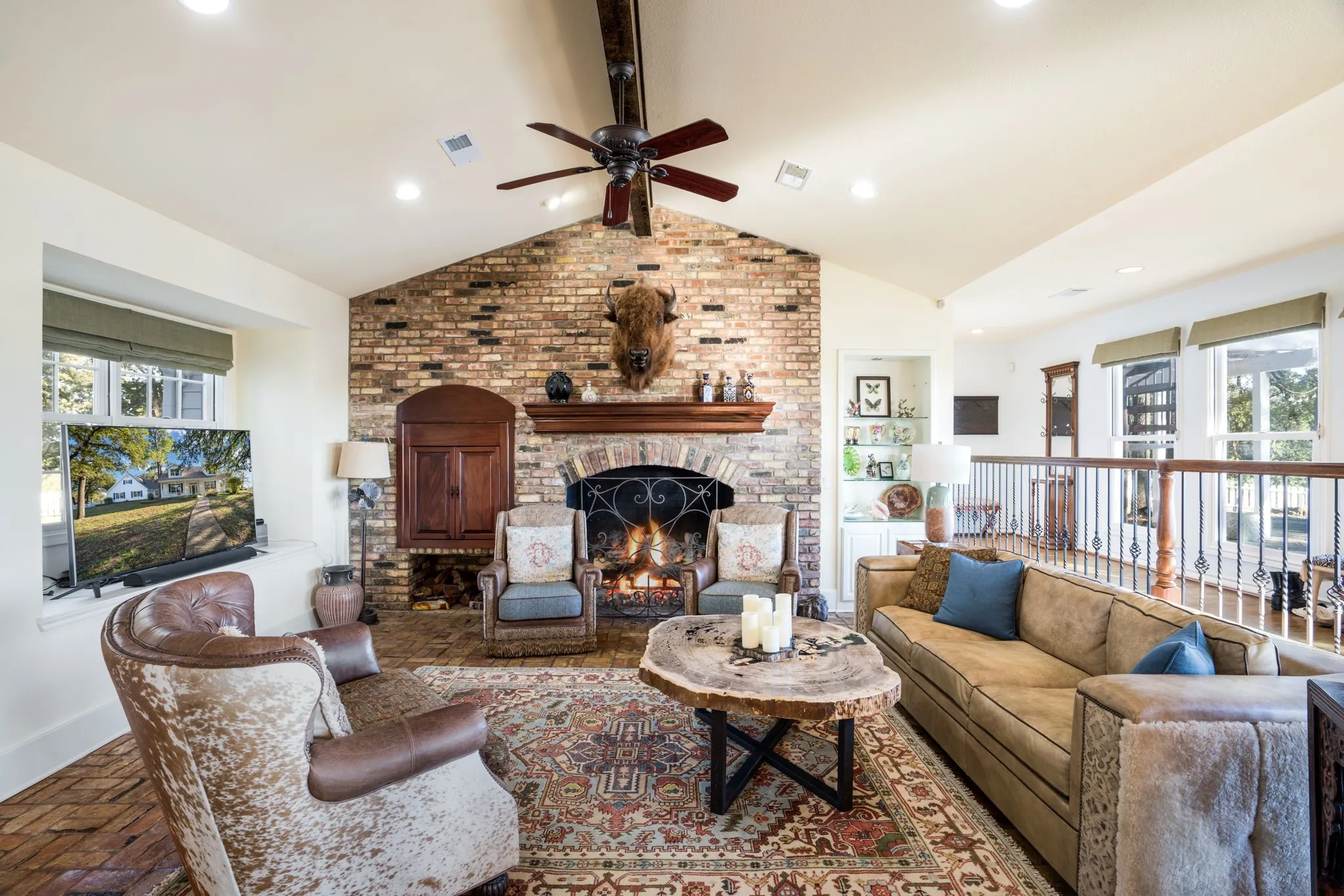Exposed brick fireplace with a wood mantle, vaulted ceiling with a ceiling fan, recessed lighting, built-in cabinetry, and a wrought iron railing