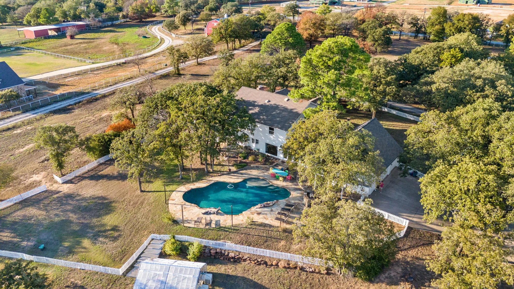 Expansive property featuring a freeform swimming pool, surrounding stone hardscaping, mature trees, and a white picket fence