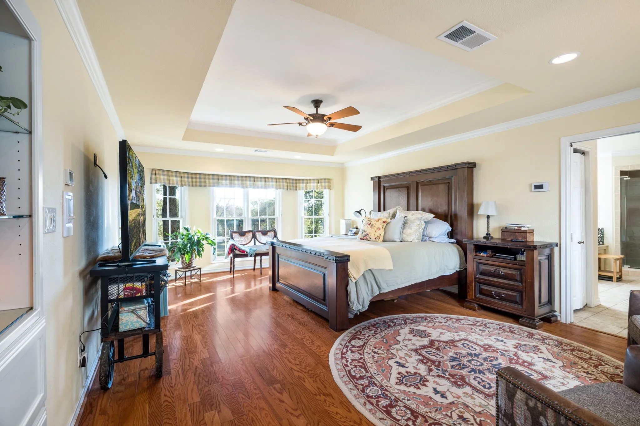 Wood-finish flooring, tray ceiling with ceiling fan, crown molding, multiple windows, and a doorway opening to an additional space
