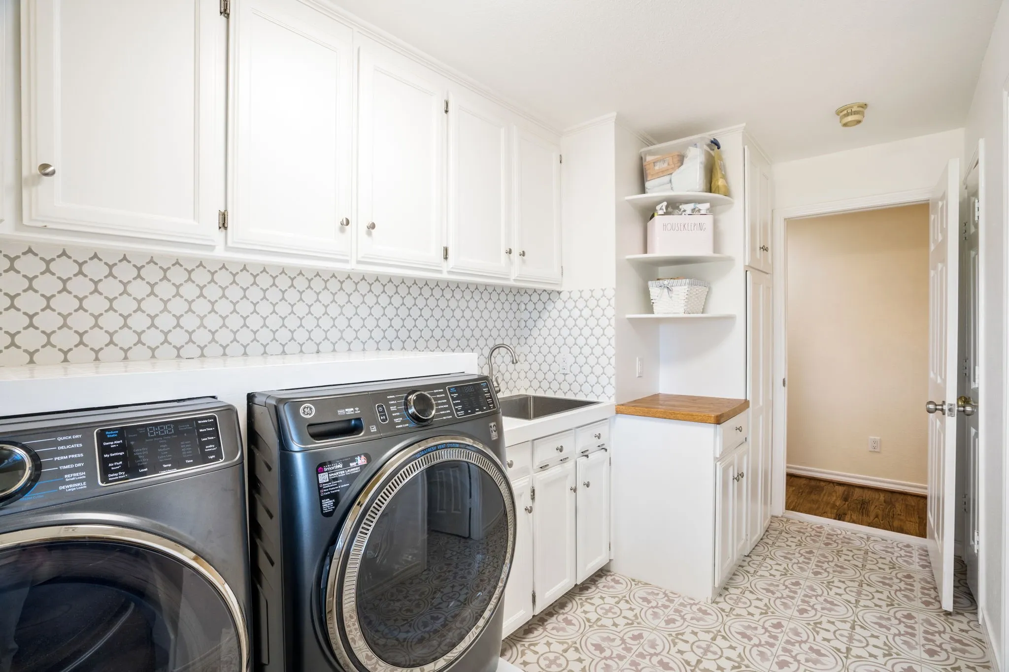 Modern laundry room featuring stainless steel front-load washer and dryer, white cabinetry with hardware accents, geometric tile backsplash, undermount sink with chrome fixtures, wood-finish countertop, and built-in open shelving