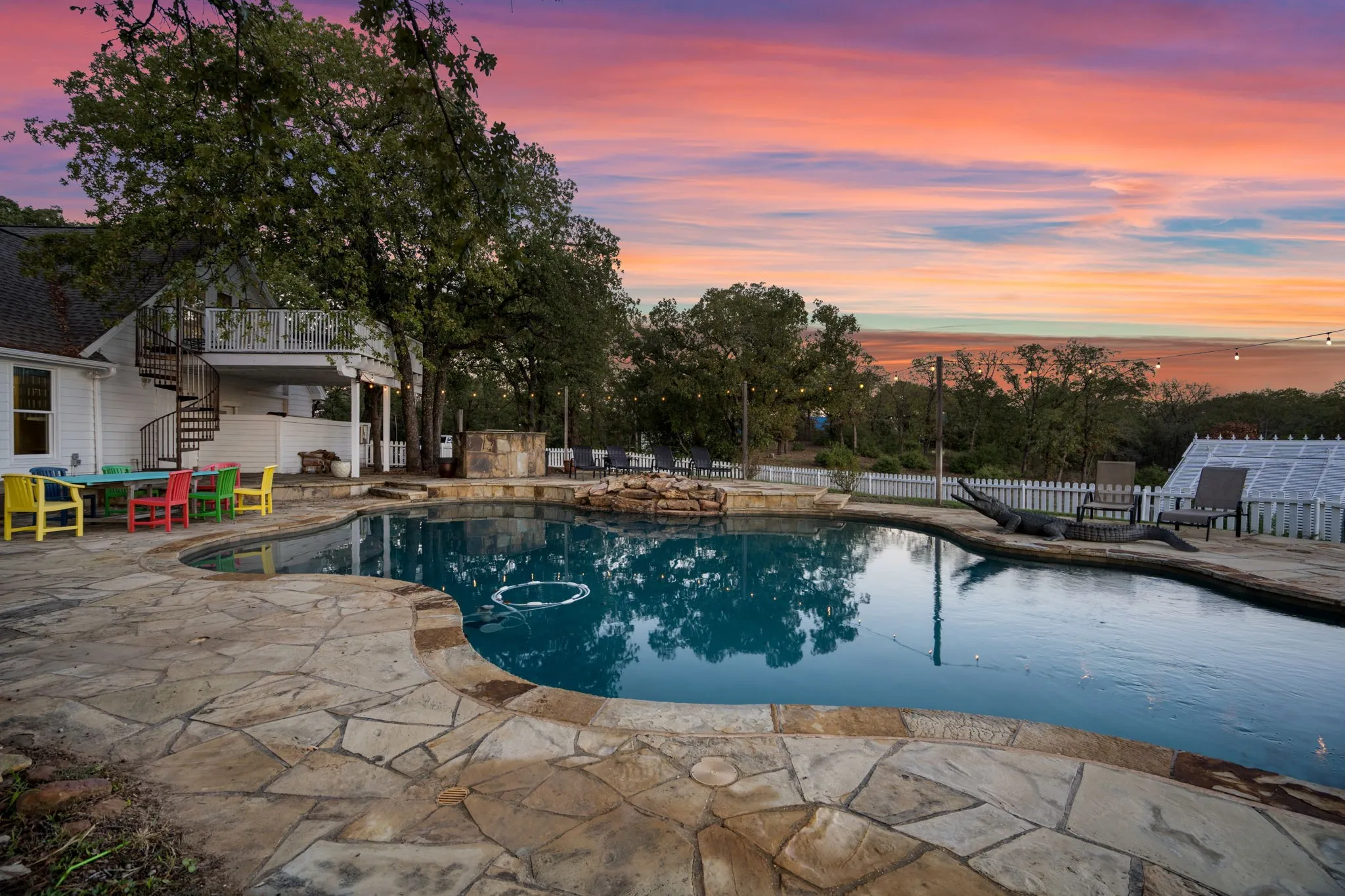 Curved swimming pool with flagstone surround, white picket fencing, and a spiral staircase leading to an upper-level balcony