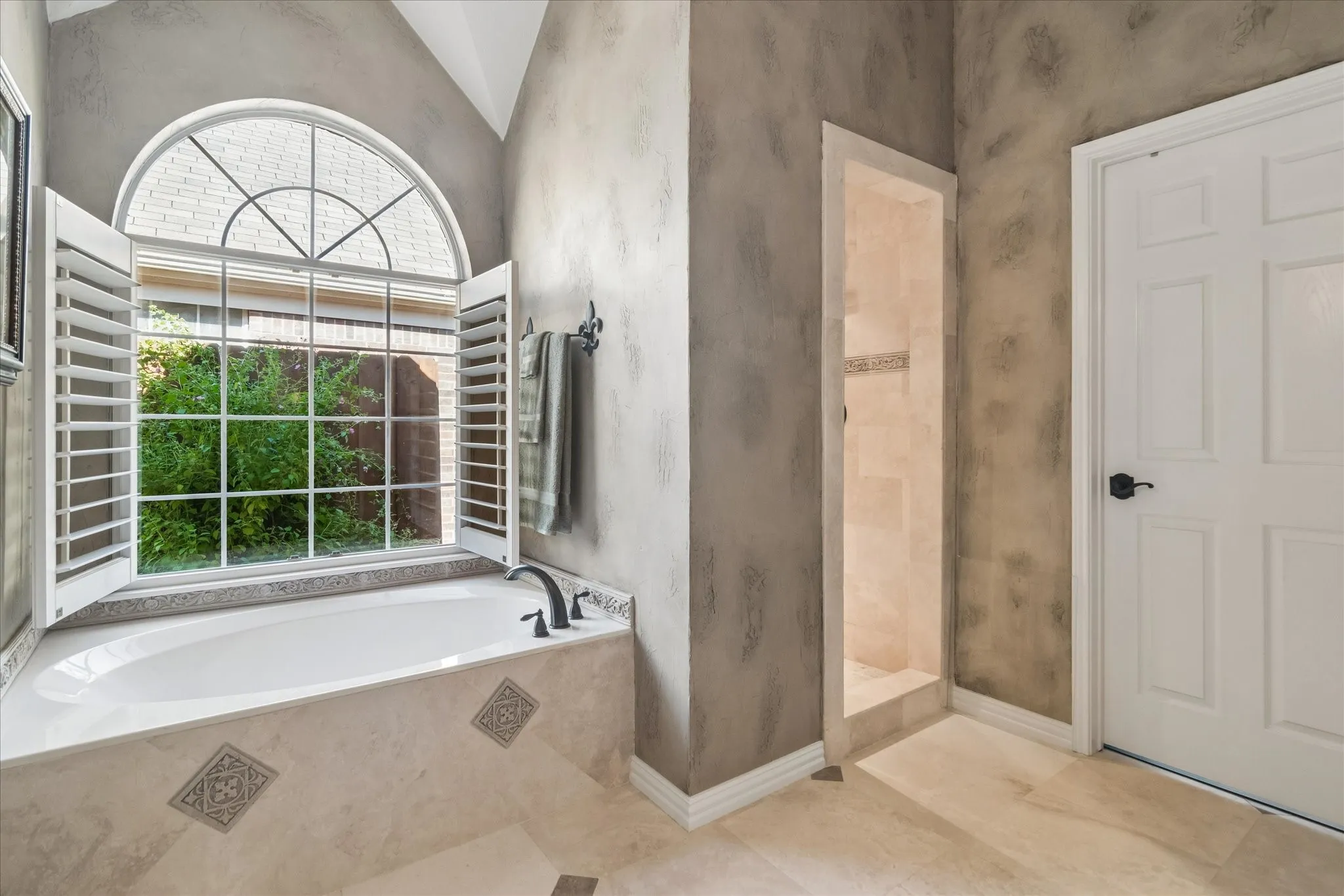Soaking tub with a black faucet, large arched window, window shutters, tiled shower, and a paneled door