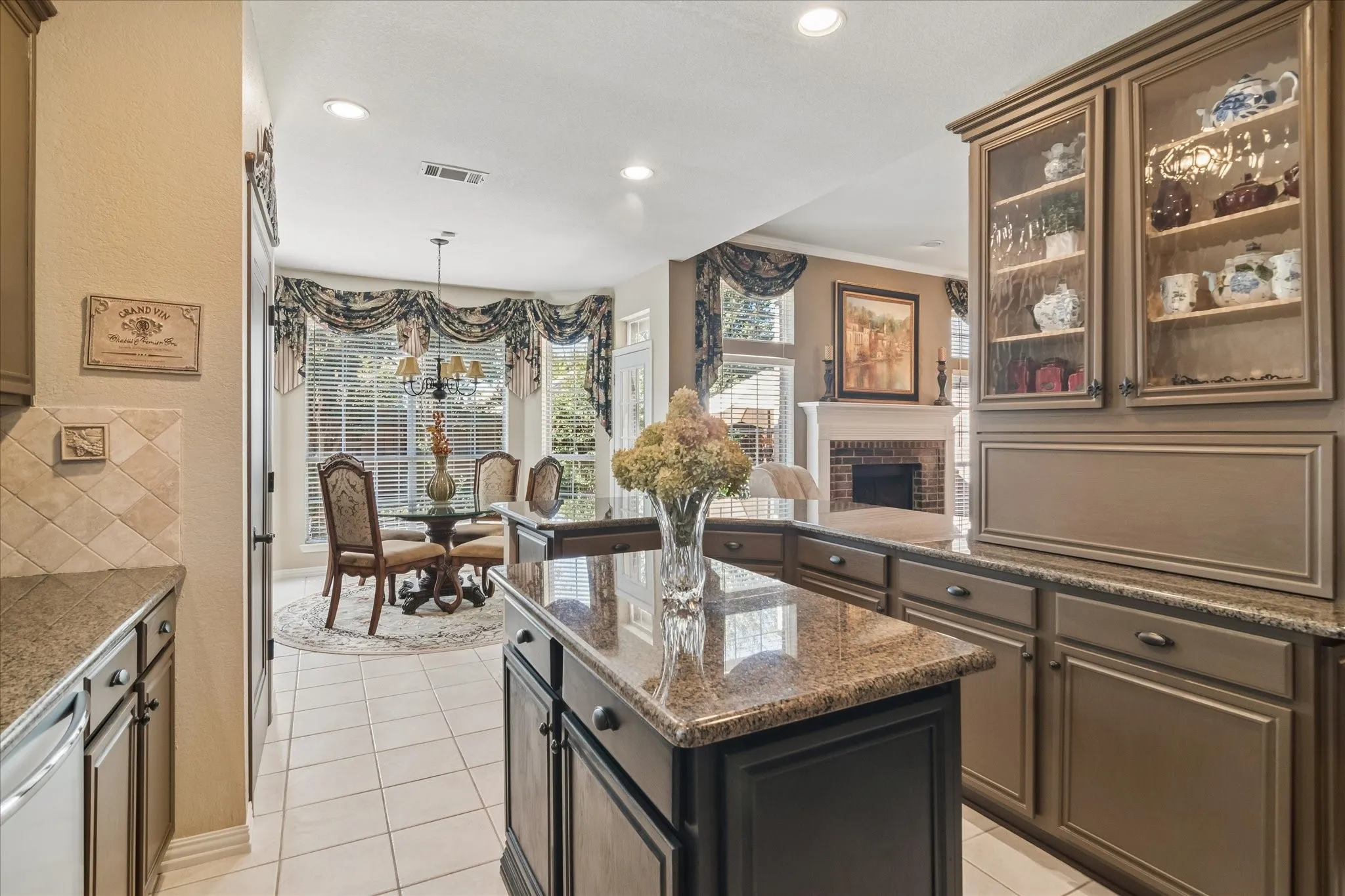 Kitchen island with a polished stone countertop, kitchen cabinetry with glass-front display cabinets, tiled flooring, an adjacent dining area with bay windows, and a brick fireplace visible in the background