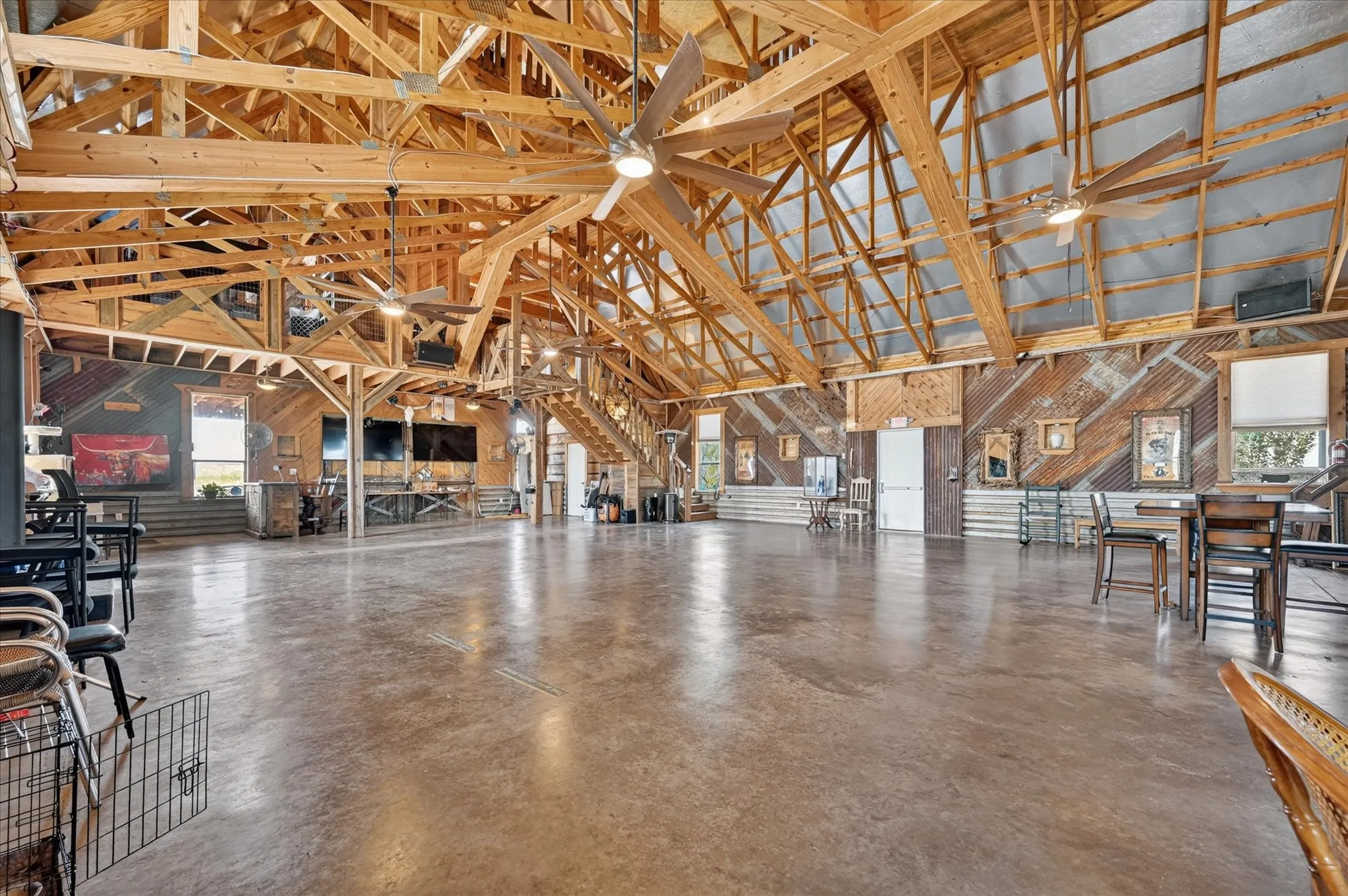 Expansive open space with high exposed beam ceilings, polished concrete flooring, multiple ceiling fans, and a visible staircase