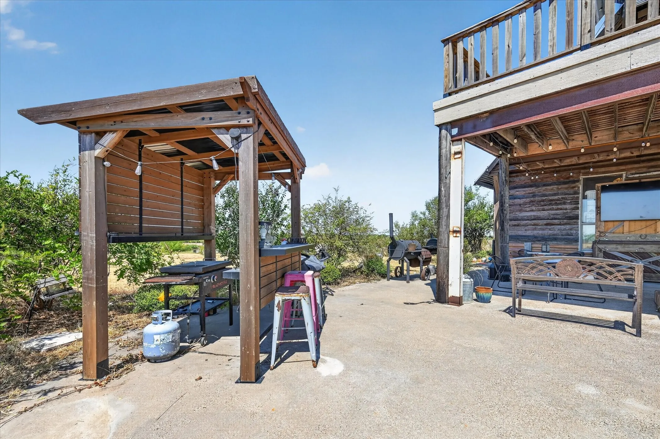 Outdoor entertainment area featuring a wood-frame overhead structure, concrete patio, and upper-level deck with wood railing