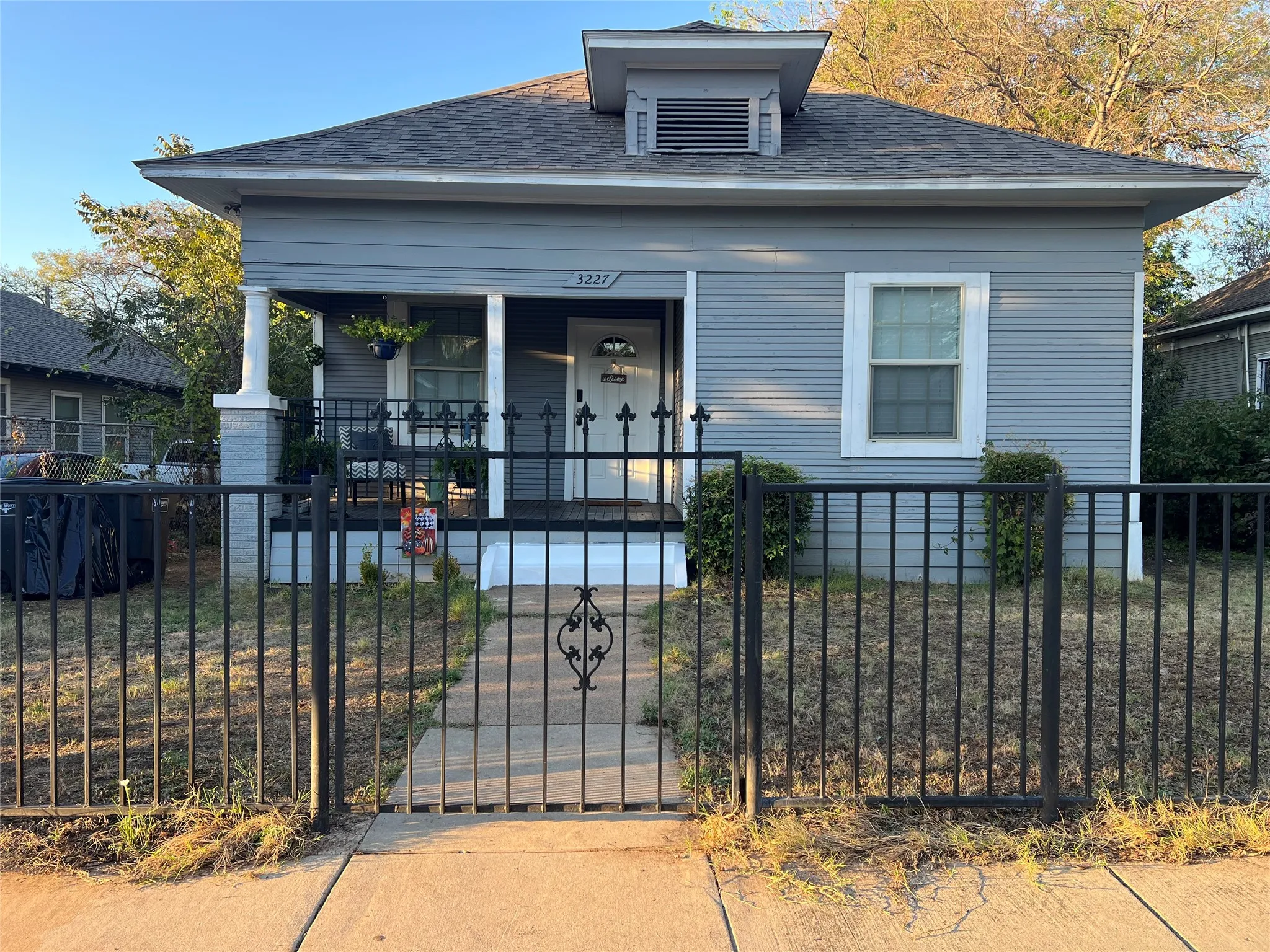 Exterior featuring light blue siding, a covered porch with white columns, a dormer with a vent, and a black wrought iron fence with an ornamental gate