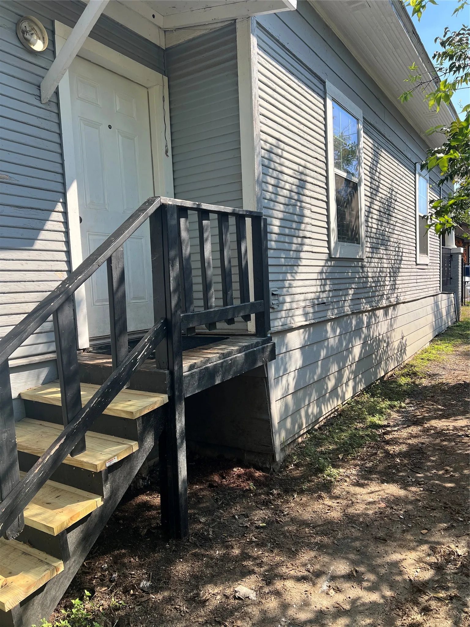 White paneled side door with overhang, light gray horizontal siding, and dark-stained wooden porch steps and railing to the kitchen.