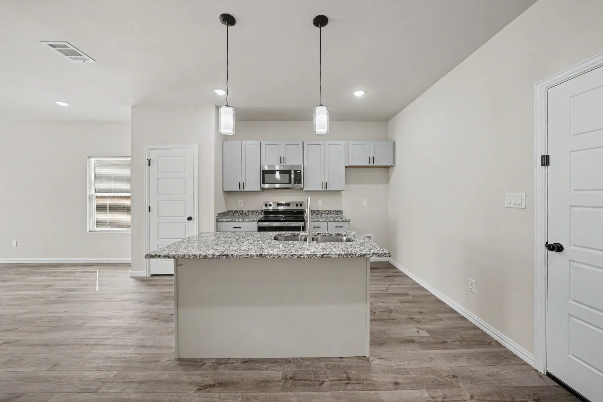 Kitchen featuring an island with a built-in sink, pendant lighting, light gray cabinetry, stainless steel appliances, and wood-finish flooring