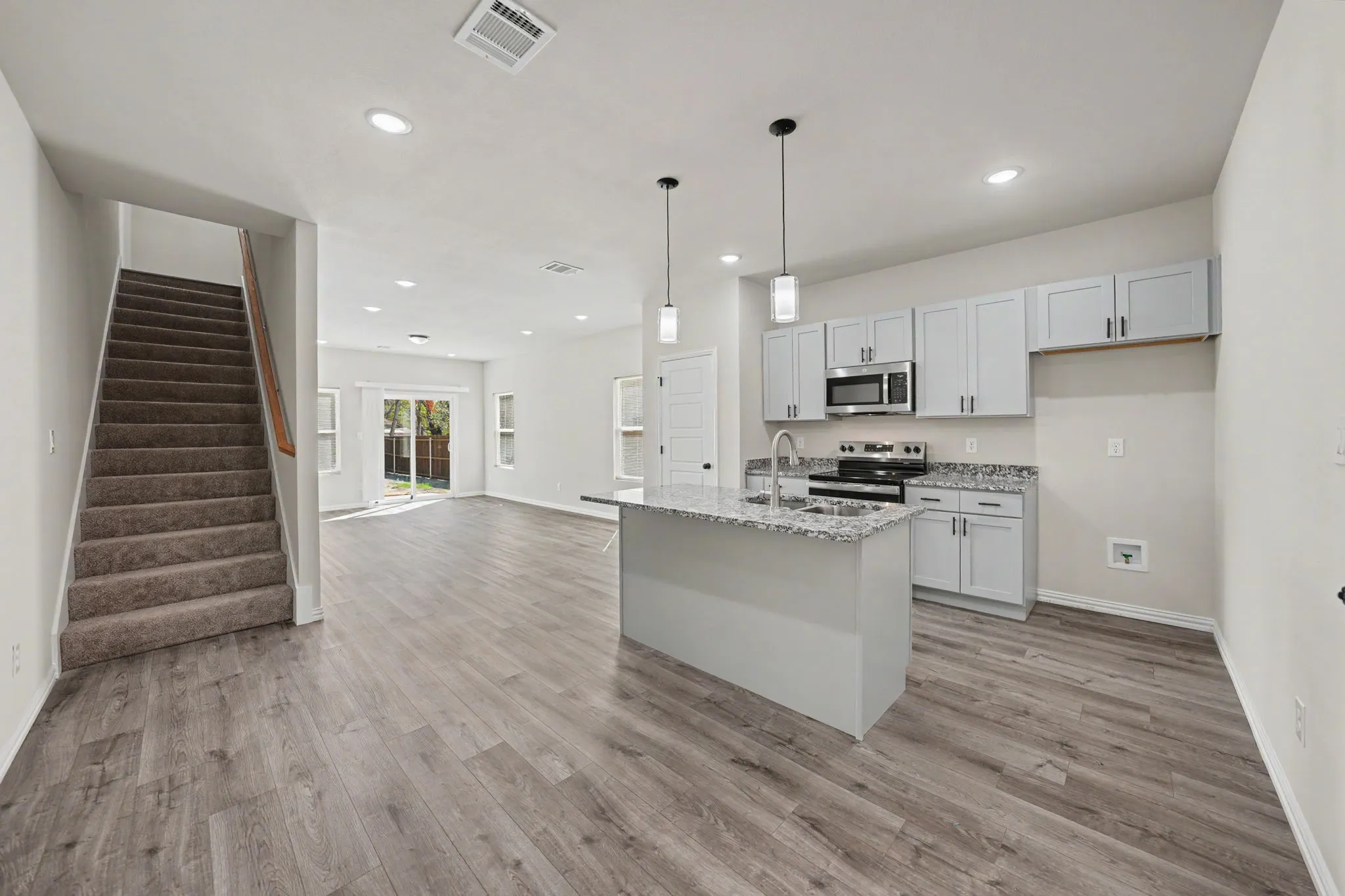 Open-concept design featuring wood-finish flooring, a kitchen island with pendant lighting, light gray cabinetry, stainless steel appliances, and a carpeted staircase