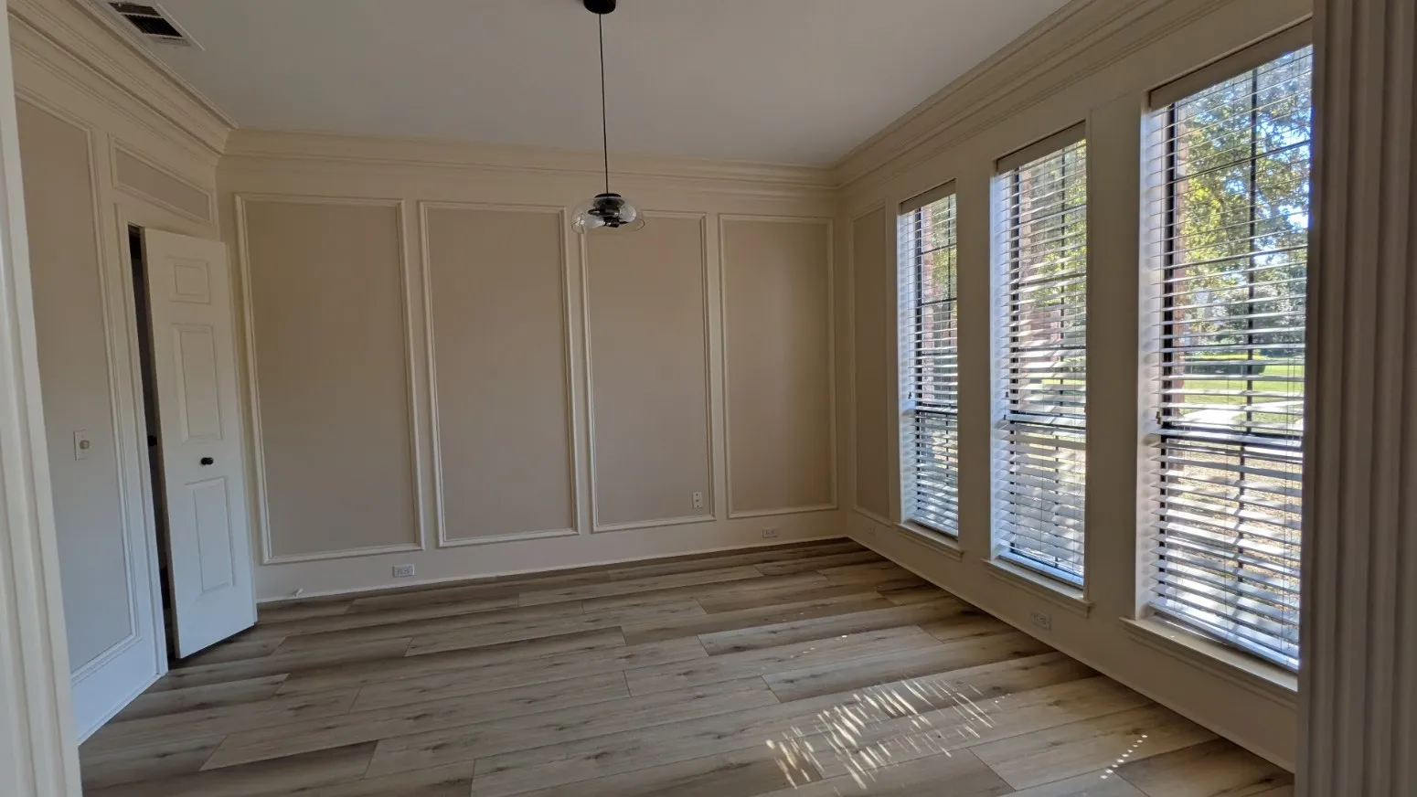Wood-finish flooring, decorative wall paneling, crown molding, three large windows, and a modern pendant light fixture