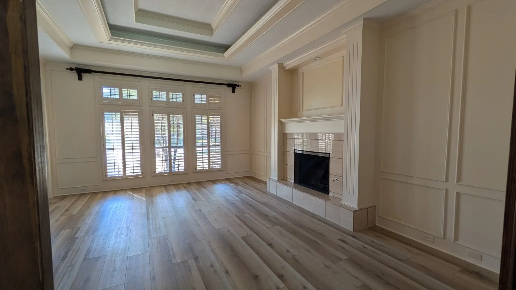 Recessed tray ceiling, wide plank wood-finish flooring, plantation shutters, tiled fireplace with mantel, and decorative wall paneling