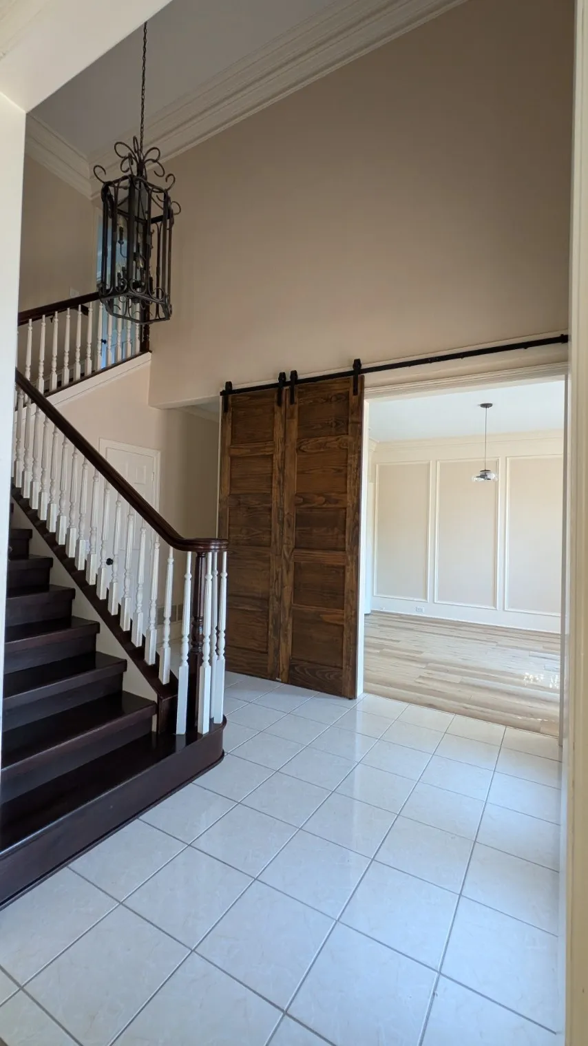 Two-story foyer featuring a dark wood-finish staircase with white balusters, square tile flooring, a decorative iron chandelier, and wood-finish barn doors leading to an additional room with a pendant light