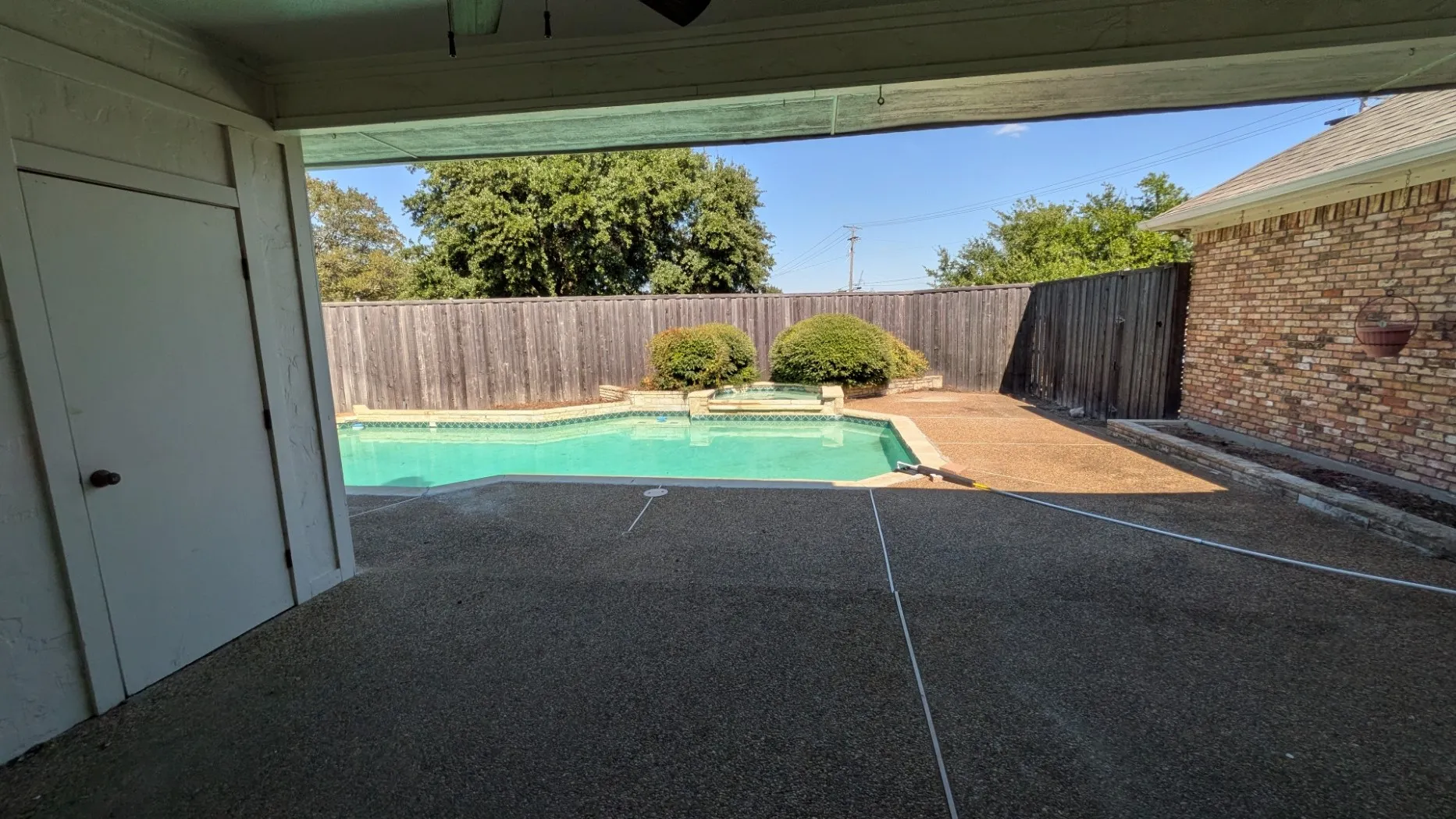 Outdoor covered patio with exposed aggregate surface, built-in swimming pool, surrounding wood fencing, and brick exterior wall
