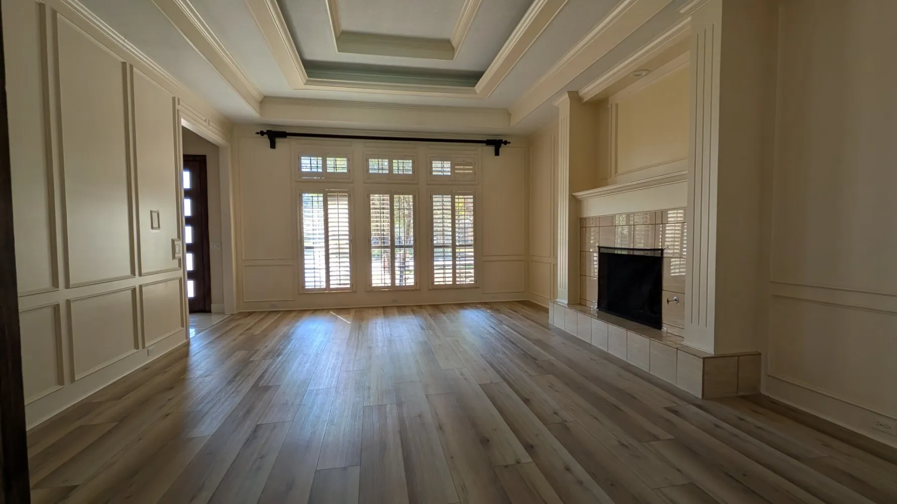 Wood-finish flooring throughout, tray ceiling with recessed lighting, decorative wall paneling, and a tiled fireplace with mantel