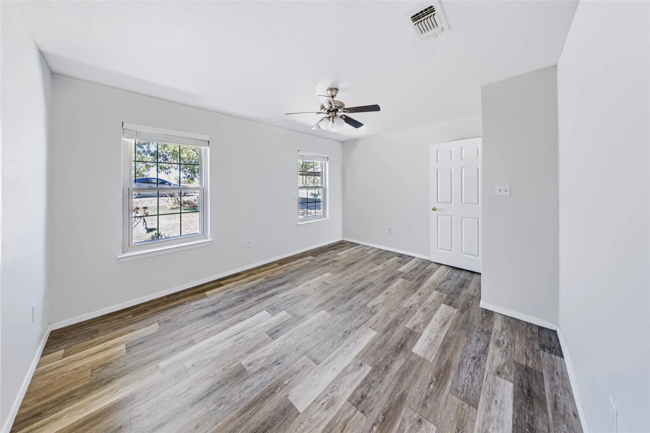 Main home — second bedroom. Wood-finish flooring, two large windows, overhead ceiling fan, and a paneled door