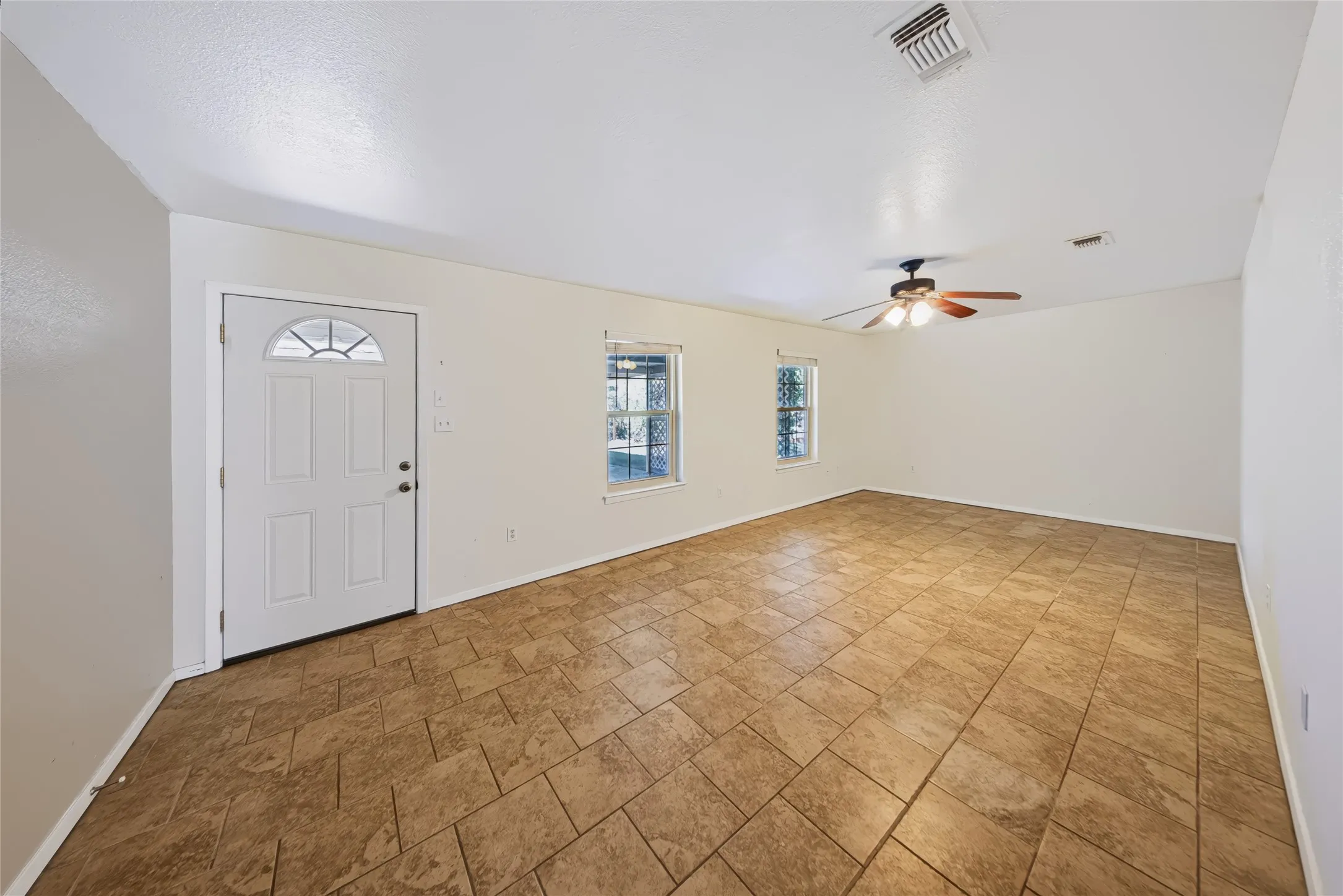 Main Home – Expansive room featuring tile flooring, a ceiling fan with integrated lighting, two windows, and a white entry door with an arched window insert