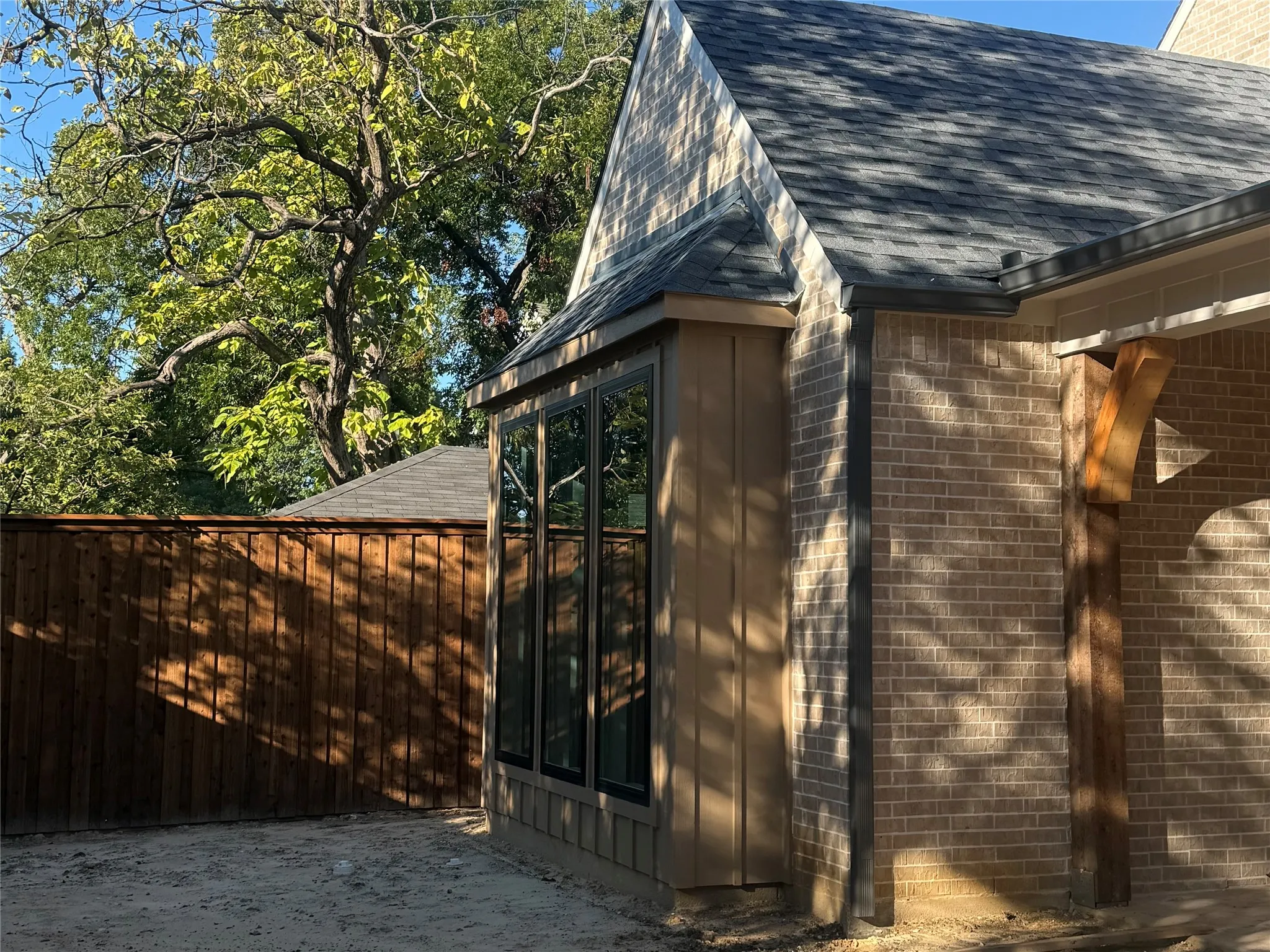 Tan brick exterior, wood siding, dark framed windows, wood beam accent, and a privacy fence