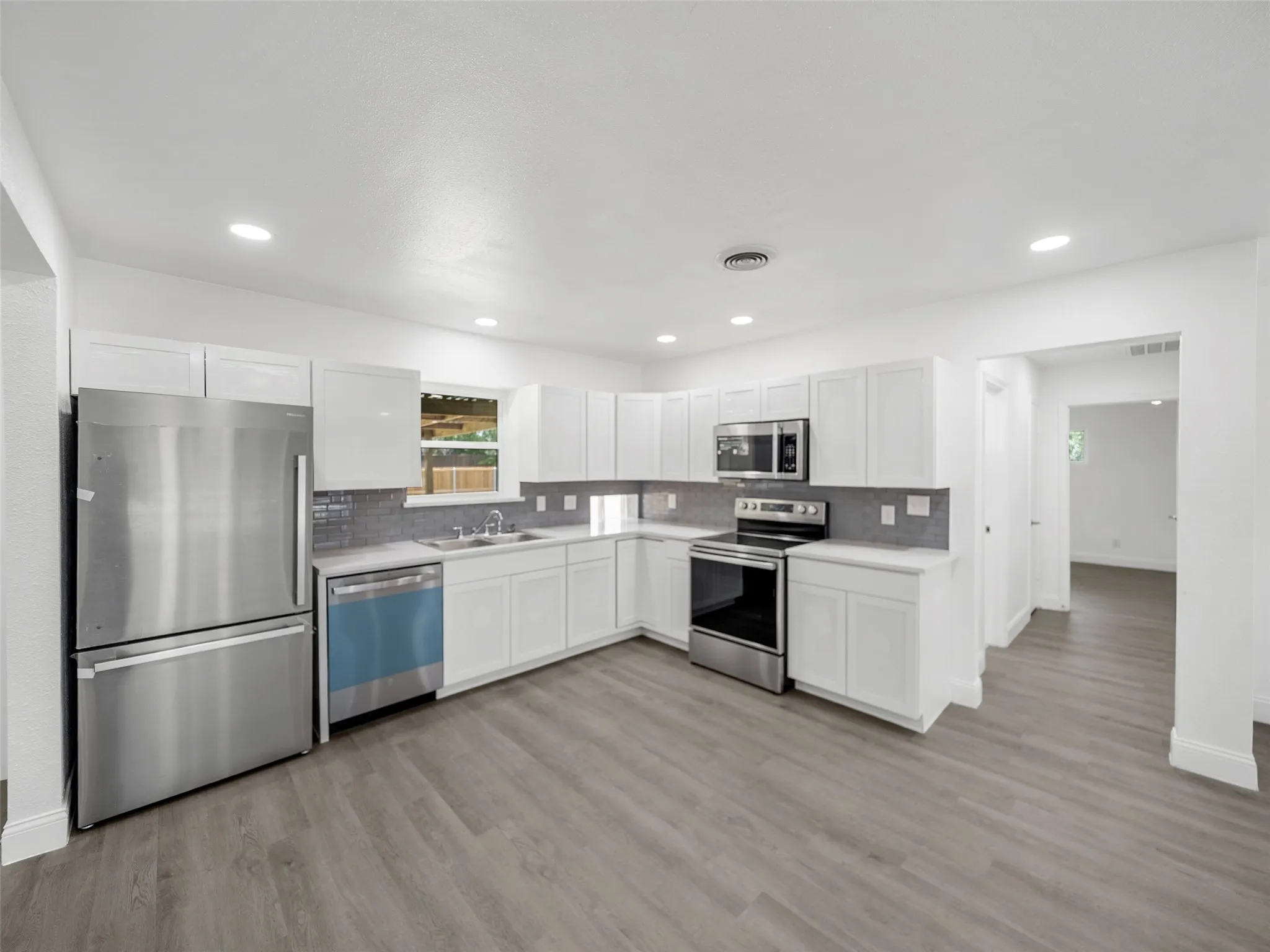 Sleek kitchen featuring stainless steel appliances, white cabinetry, gray tile backsplash, light countertops, and wood-finish flooring