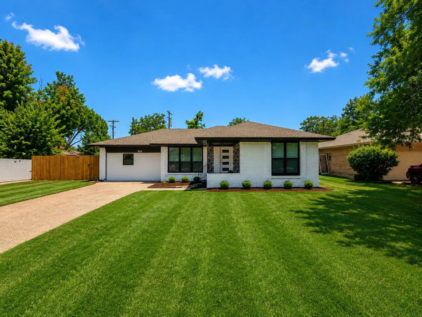 White brick exterior with dark trim windows, attached garage, gravel driveway, and a front lawn with mature landscaping