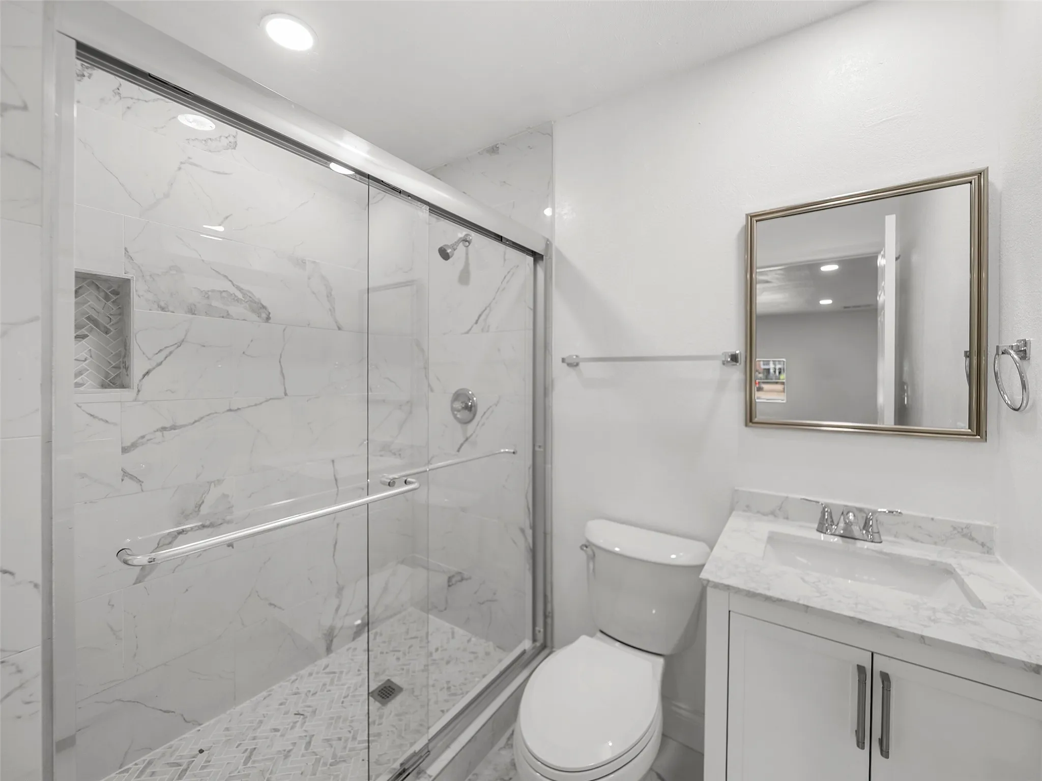 Sliding glass shower enclosure with white marbled tile surround, recessed shower niche with herringbone tile, white vanity with integrated sink and marbled countertop, and a framed vanity mirror