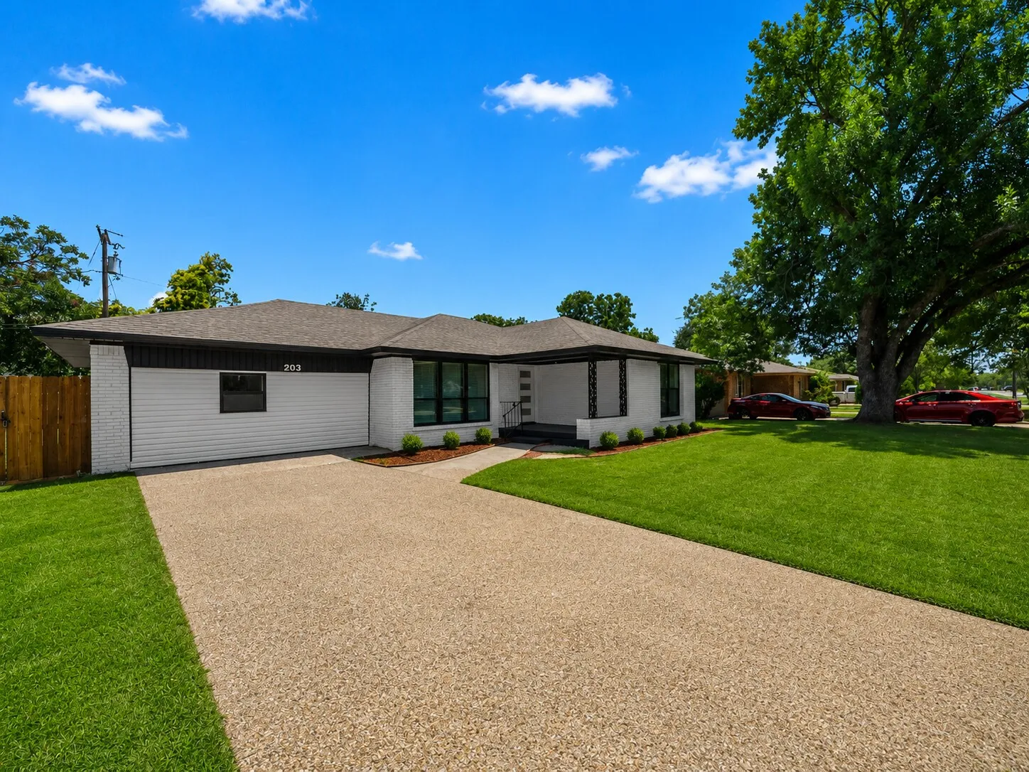 White brick exterior with black trim, gravel driveway, manicured lawn, and mature trees